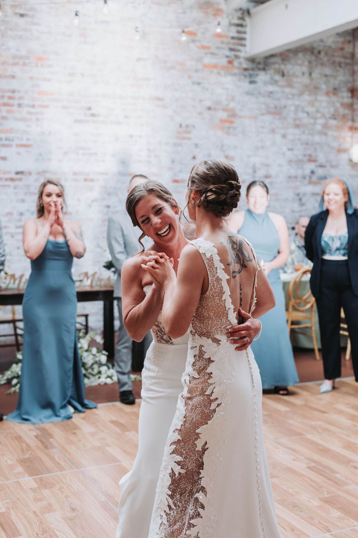 Two brides sharing their first dance while guests in blue dresses applaud at wedding reception.