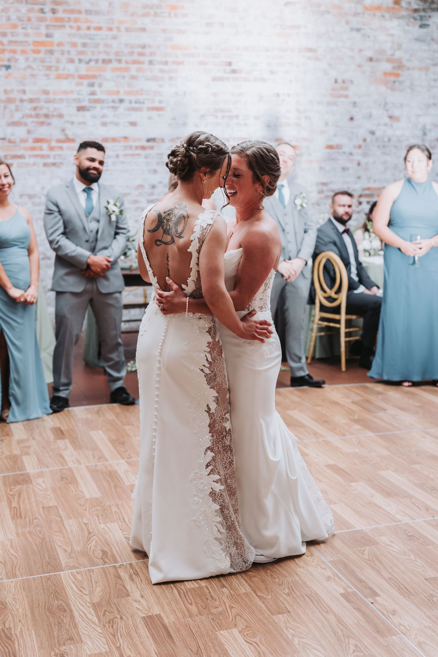 Two brides laughing and dancing together in elegant white gowns on wooden dance floor at wedding reception.
