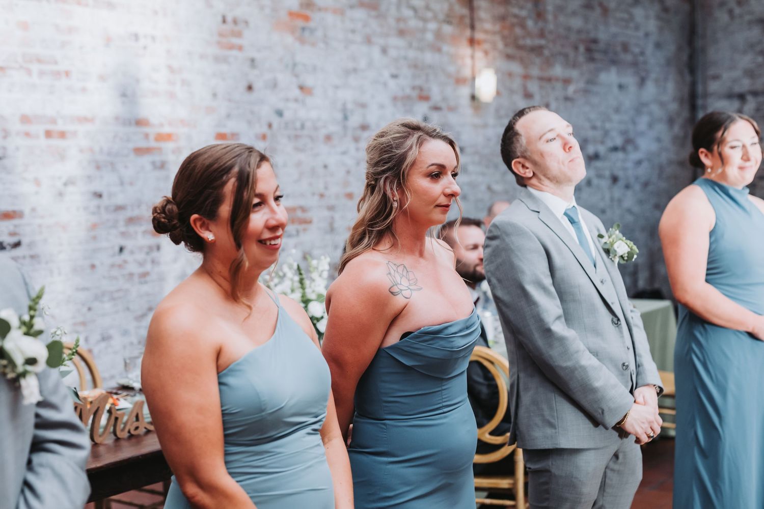 Bridesmaids in dusty blue dresses and groomsman in gray suit watching emotional wedding ceremony at brick wall venue.