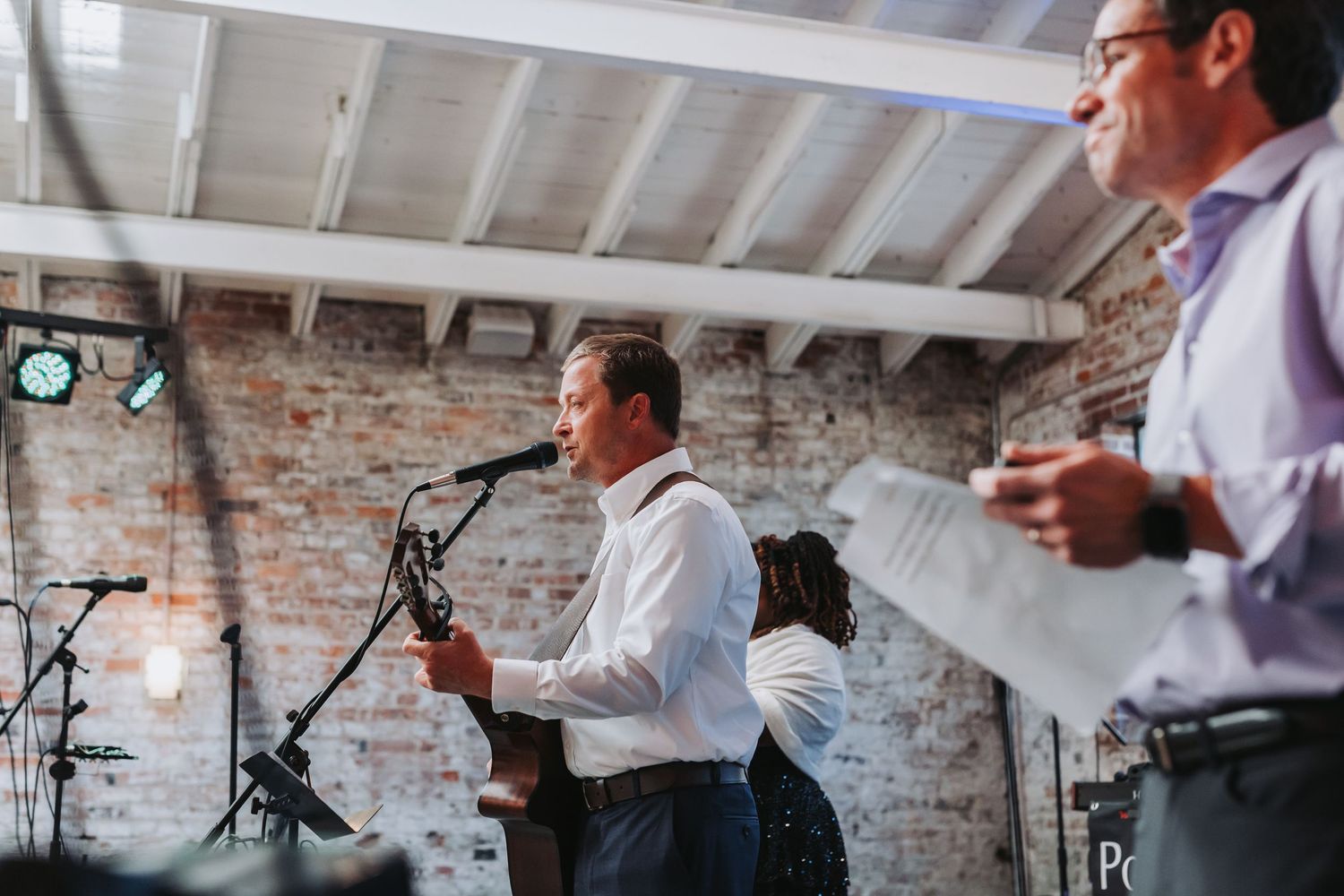 Man speaking into microphone giving wedding toast at rustic brick wall venue reception.