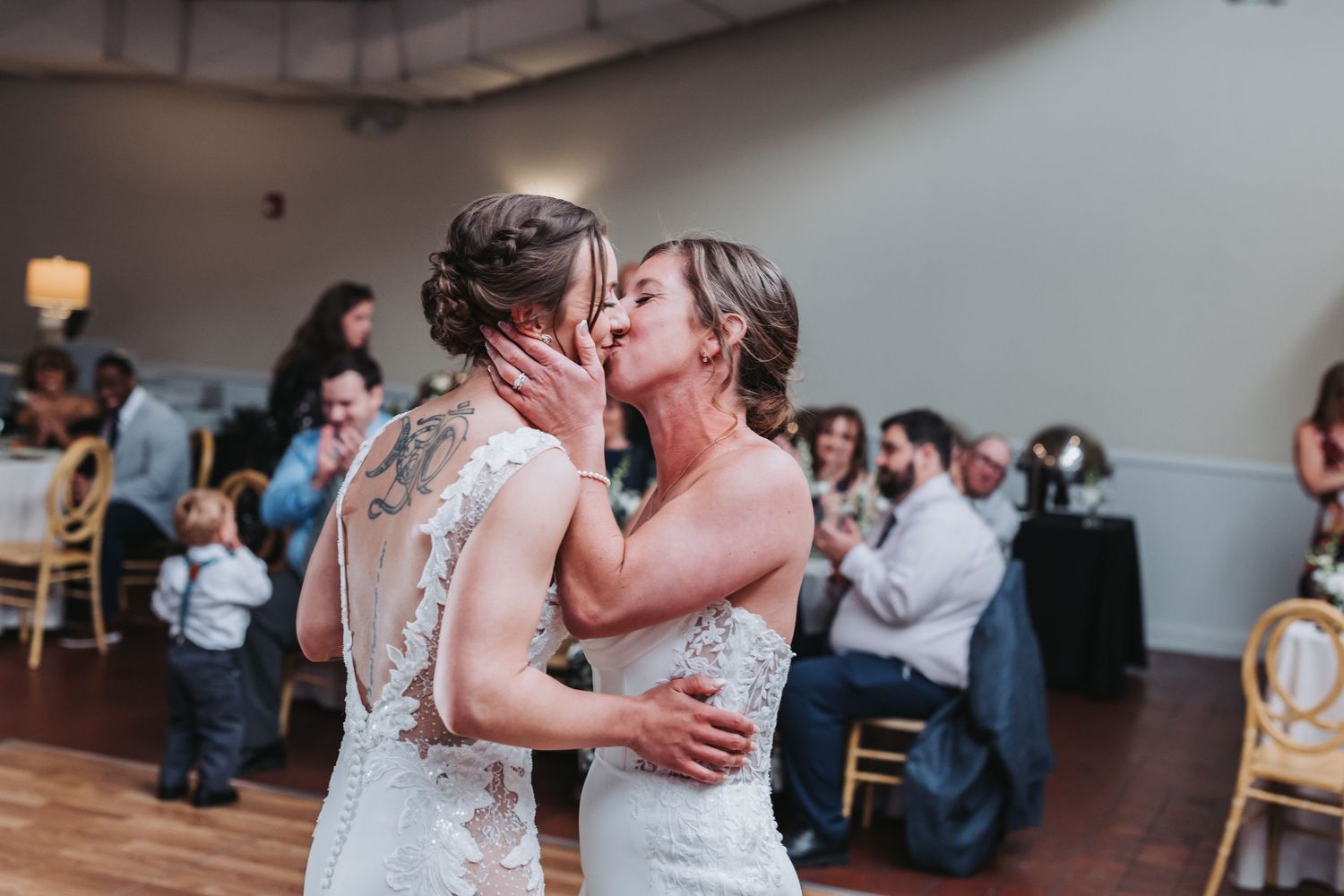 Two brides sharing a romantic kiss during their first dance at an indoor wedding reception.
