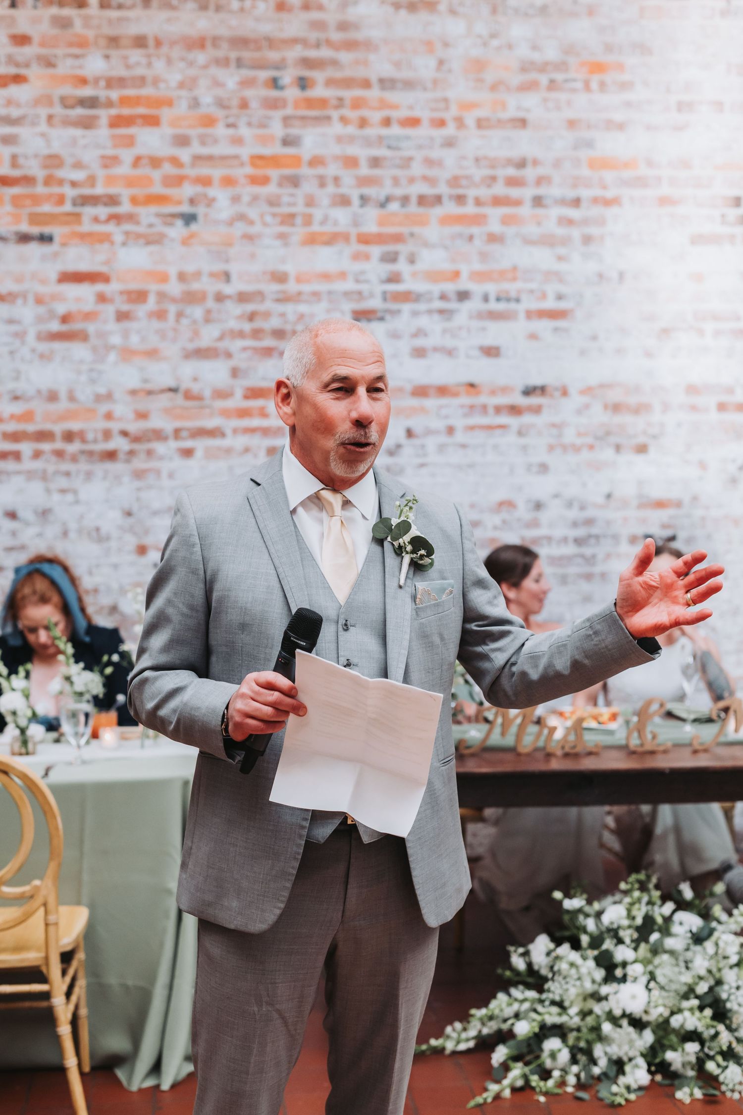 A man in a gray suit delivers a heartfelt wedding speech holding a microphone and paper before a brick wall.