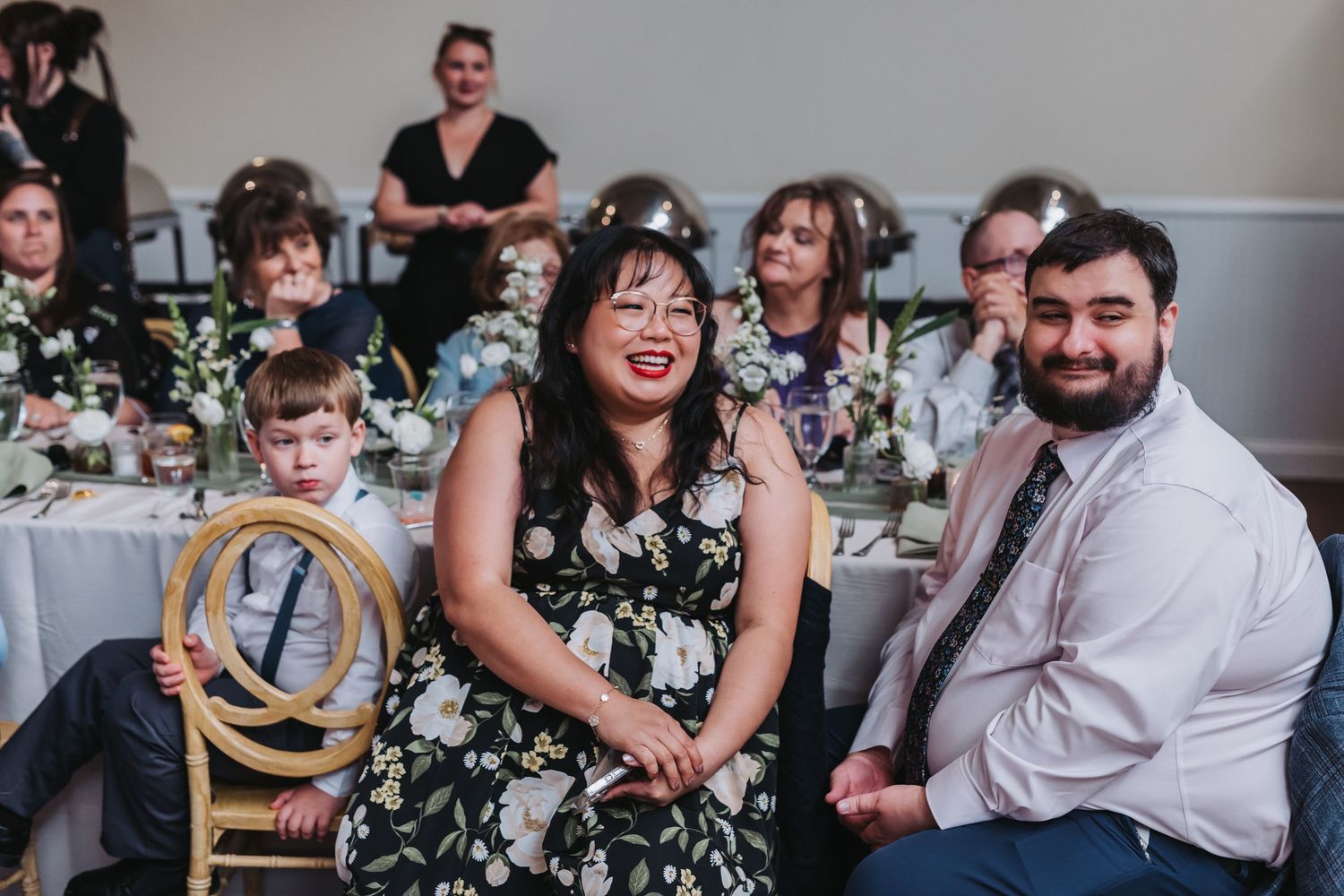 Wedding guests smiling and laughing at reception table with floral centerpieces.