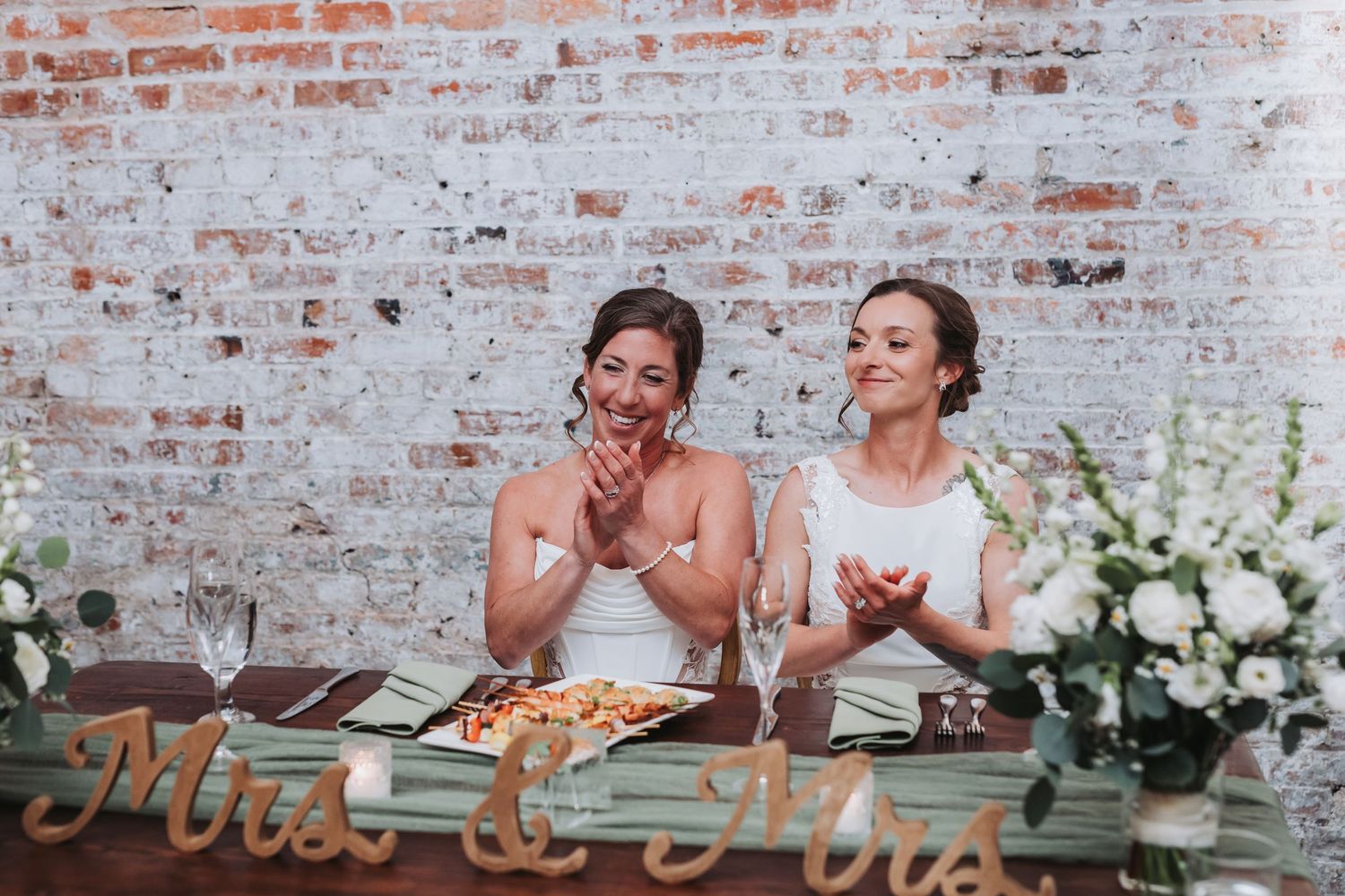 Two brides in white dresses clap joyfully at their wedding reception table with a Mrs. and Mrs. sign.