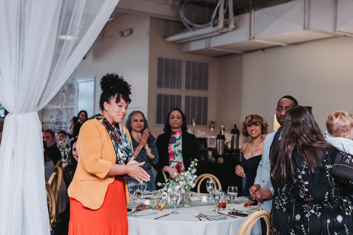 Woman in orange jacket giving a heartfelt wedding speech to seated guests at a decorated reception table.