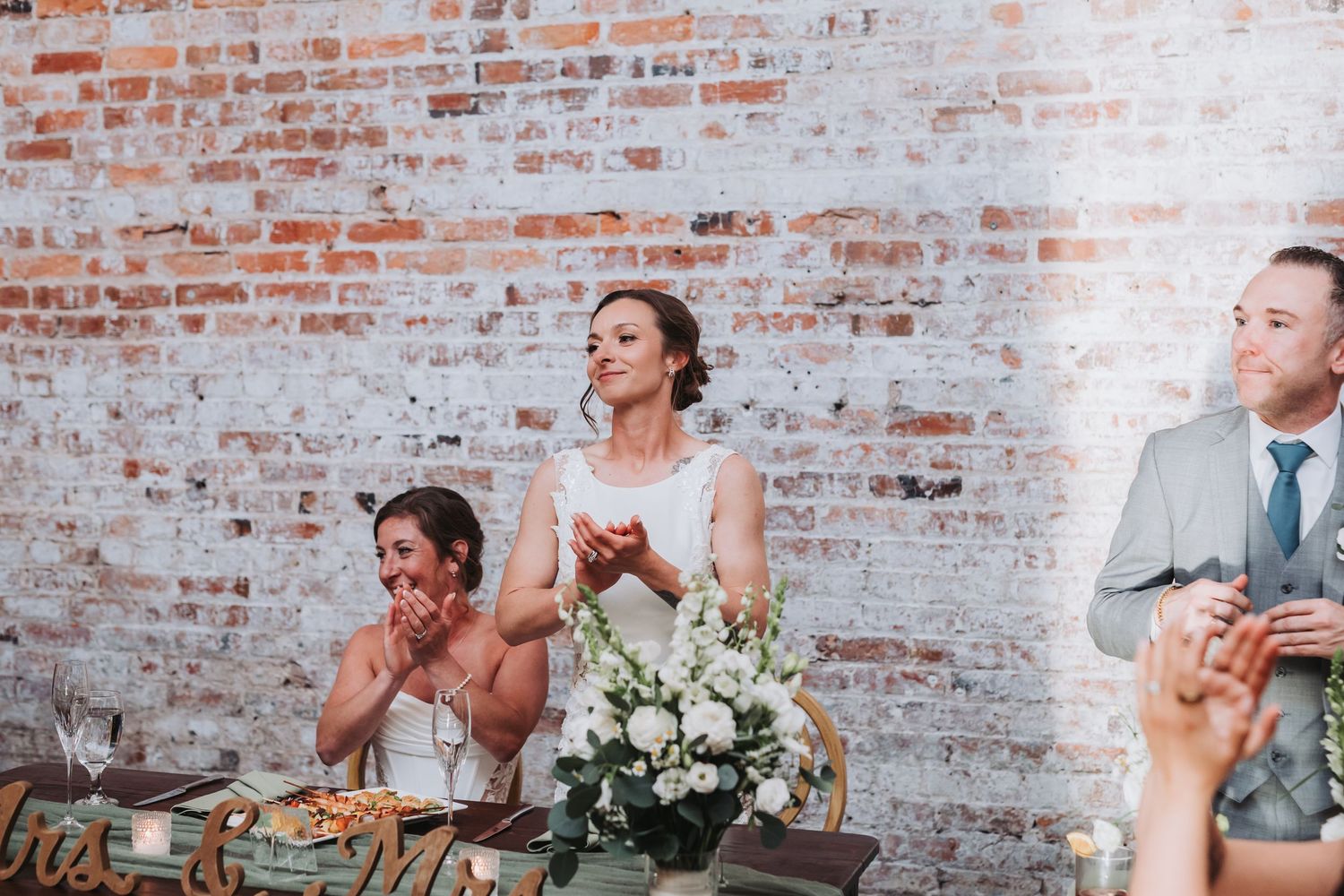 Bride in white gown clapping at wedding reception table with floral centerpiece and brick wall.