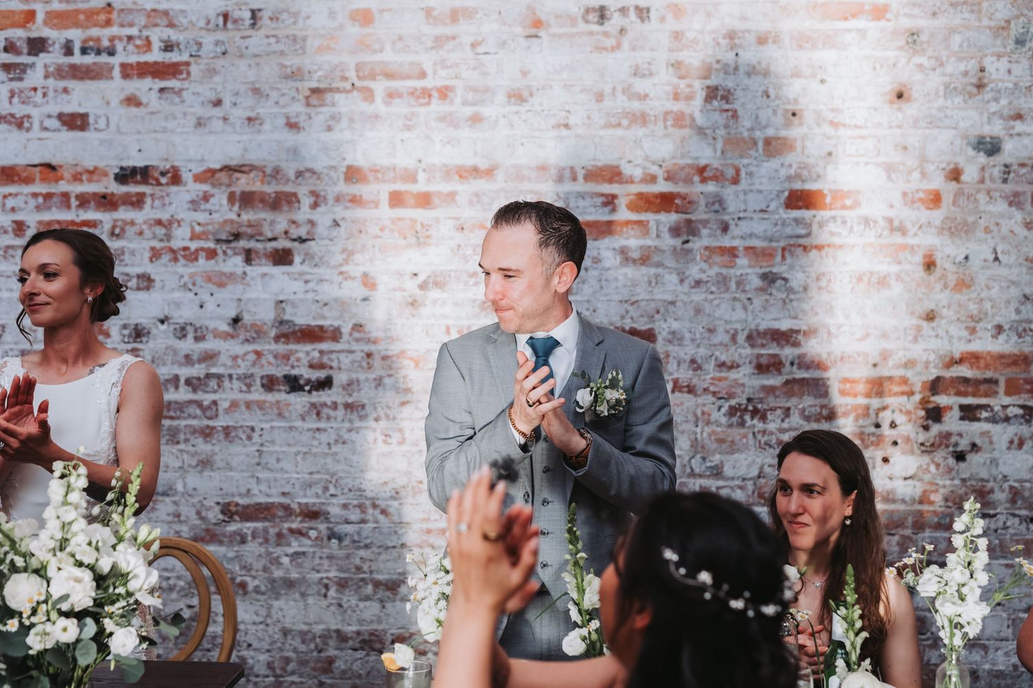 Smiling groomsman in gray suit applauding during wedding toasts at reception with exposed brick wall backdrop.