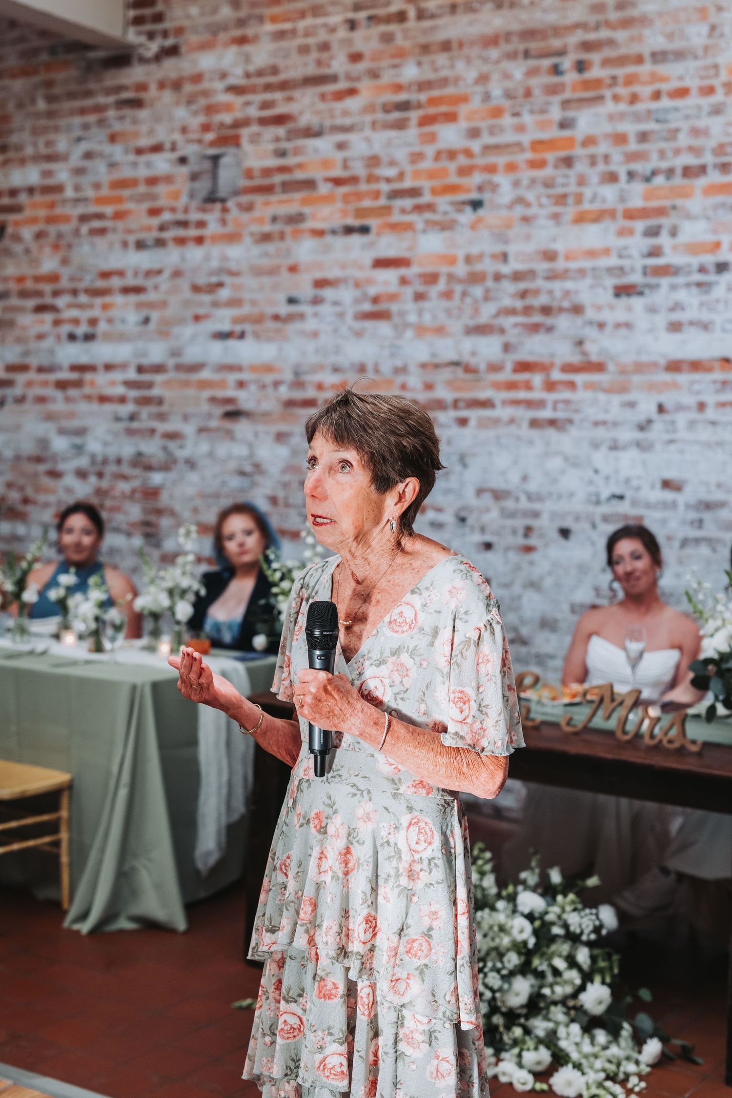 Elderly woman in floral dress giving a heartfelt speech at a wedding reception with brick wall backdrop.