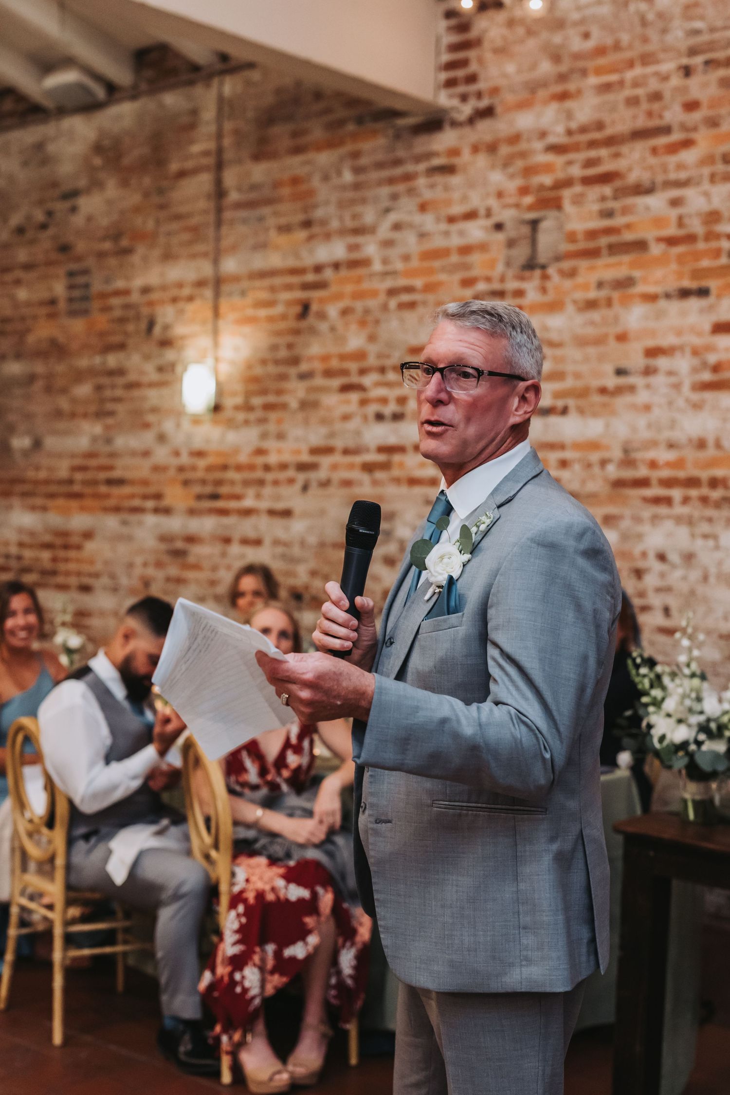 Father of the bride delivers heartfelt speech in light gray suit at wedding reception against exposed brick wall.