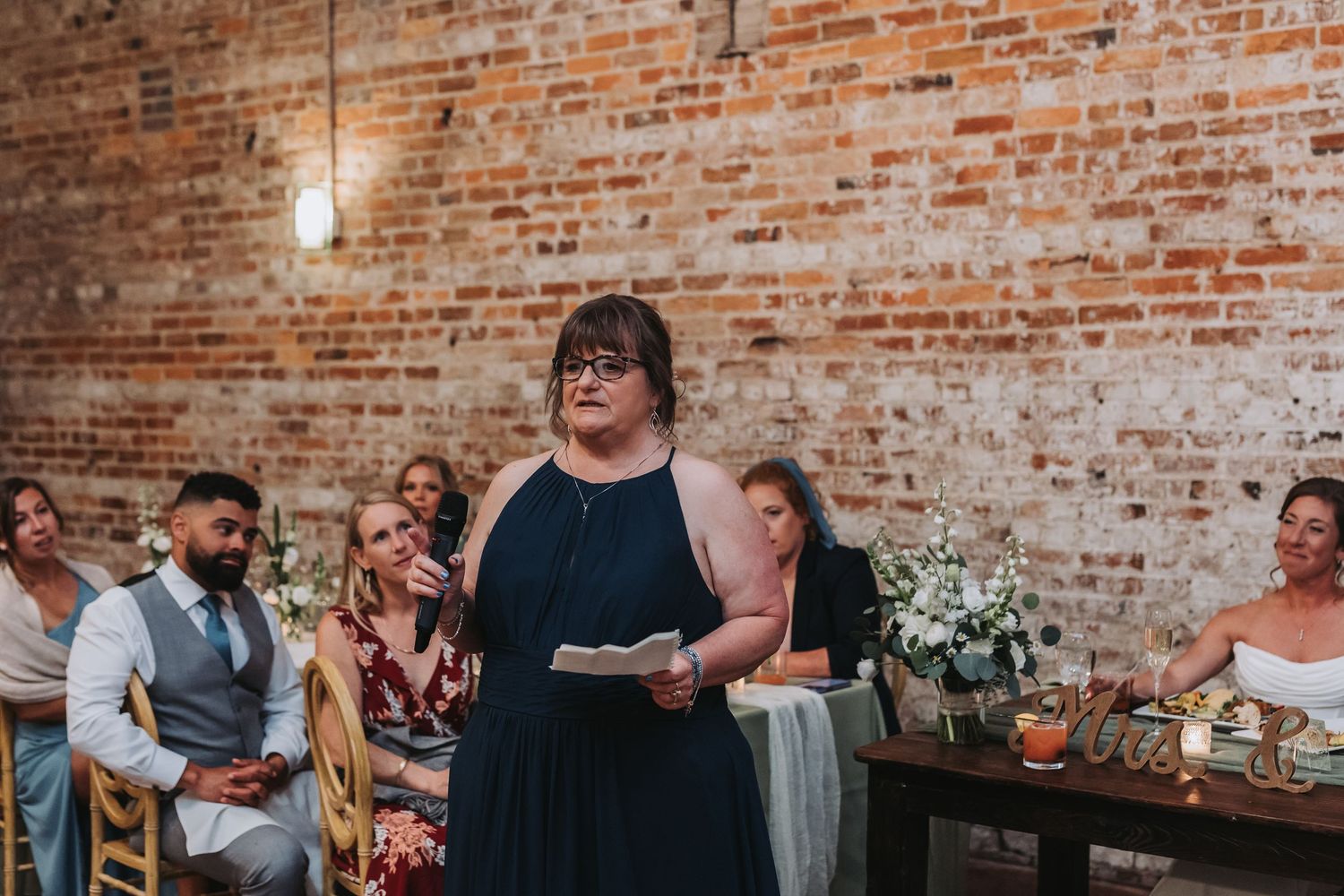 Woman in navy dress giving speech at wedding reception in rustic brick venue.