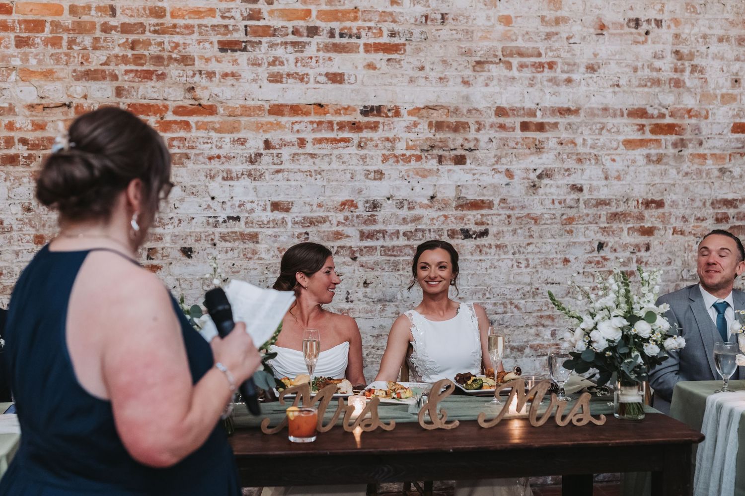 Two brides smiling at sweetheart table while guest reads heartfelt speech at rustic brick wall wedding reception.