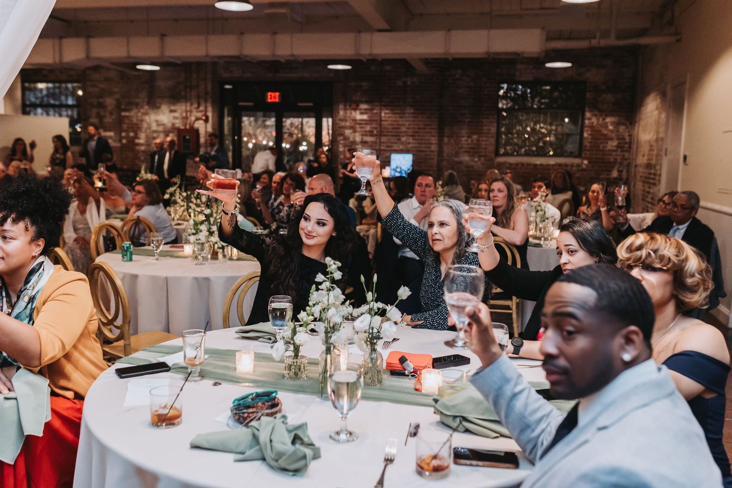 Wedding reception guests raising glasses in a toast inside a rustic brick venue with round tables.