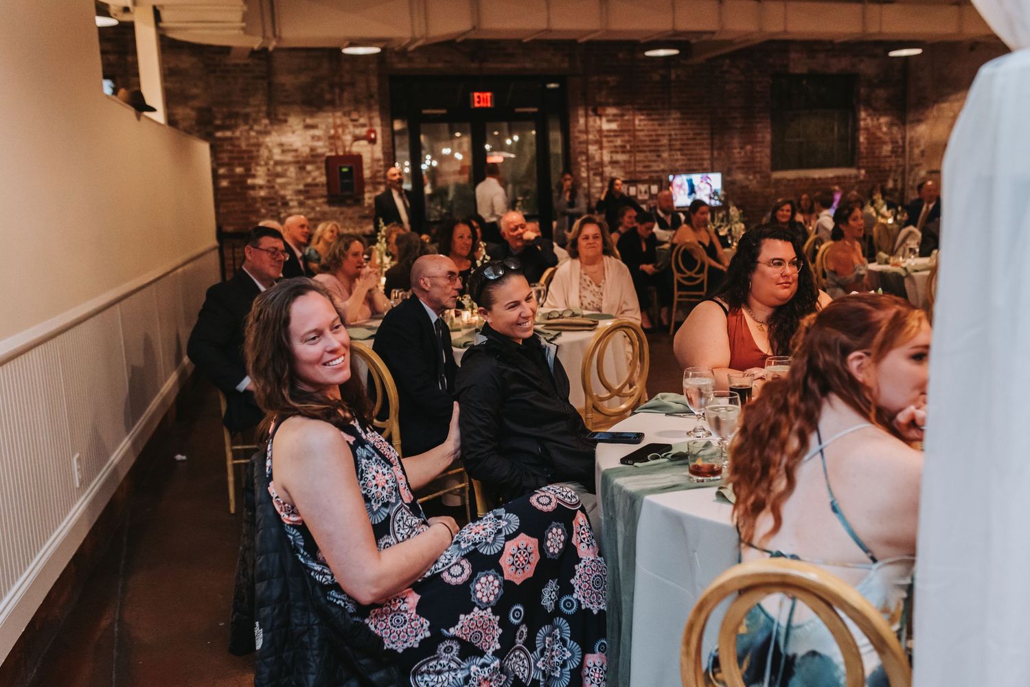 Wedding reception guests seated at decorated tables smiling and enjoying the celebration in a brick venue.