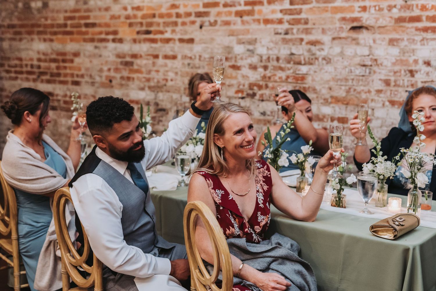Wedding guests raising glasses in a toast at a reception with brick walls and floral centerpieces.