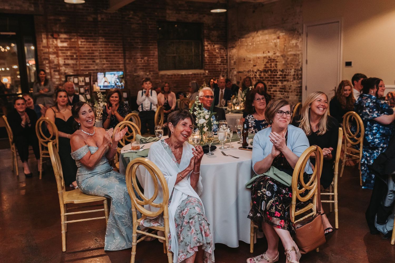 Wedding reception guests laughing and applauding during speeches in a rustic venue with exposed brick walls.