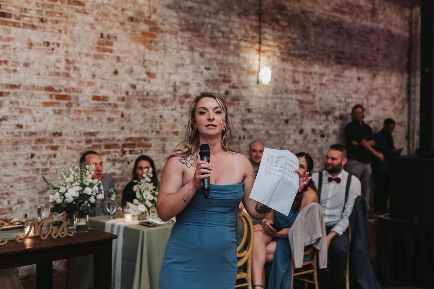 Woman in blue dress giving a speech with microphone at an indoor wedding reception.