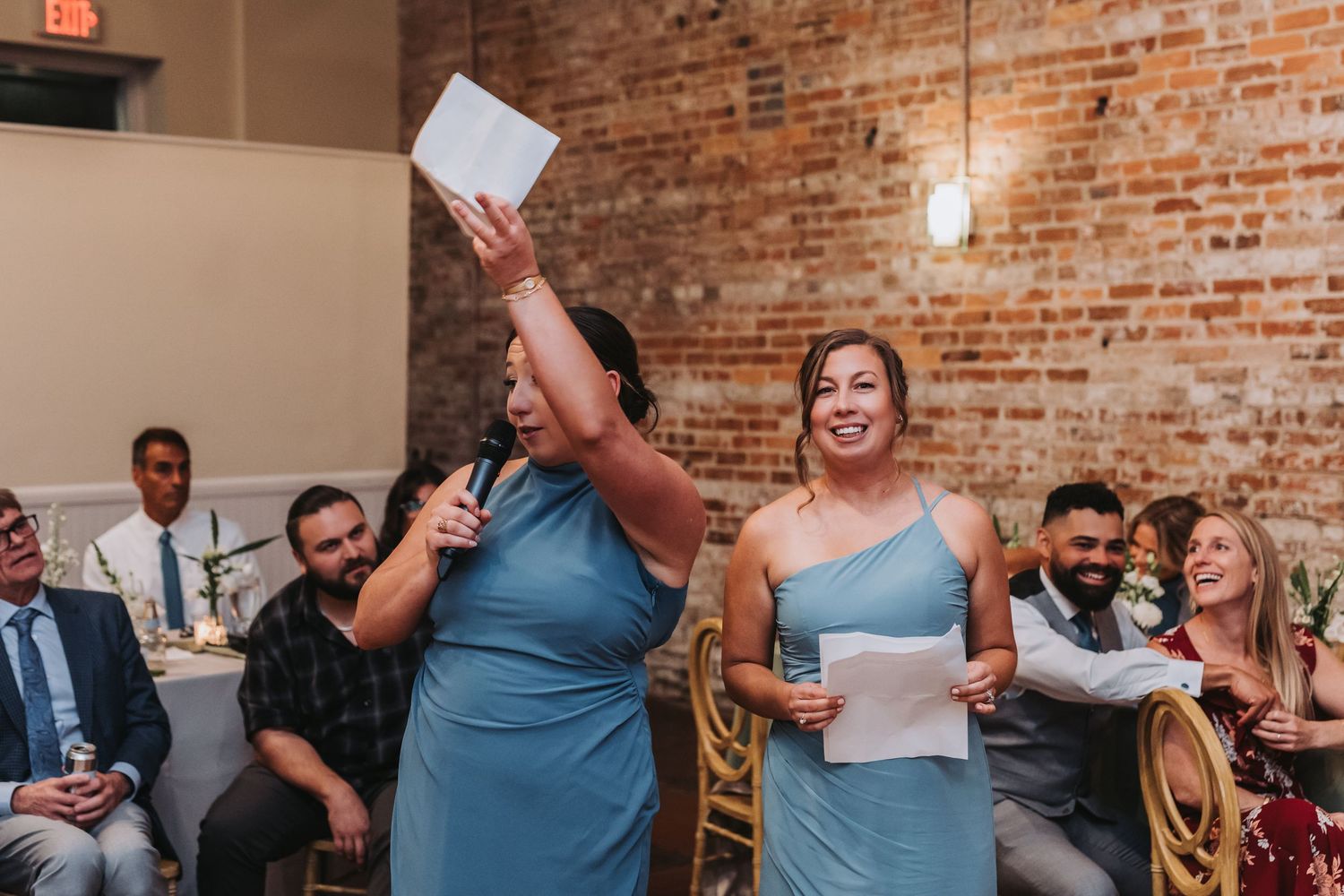 Two bridesmaids in blue dresses deliver toast speech at wedding reception in rustic brick venue.