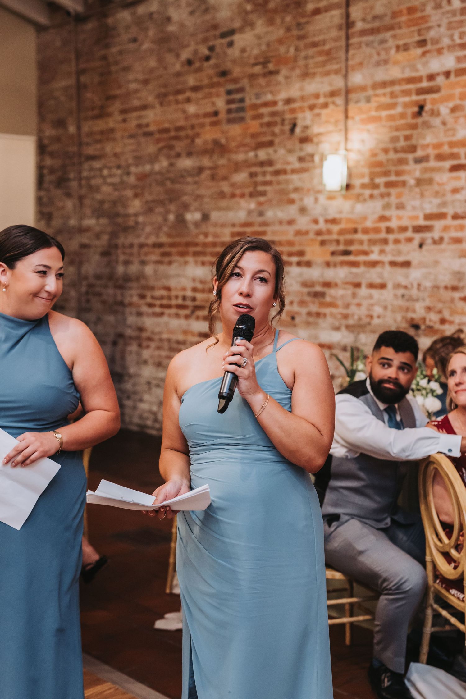 Bridesmaid in blue dress gives wedding speech with microphone at rustic brick wall venue reception.
