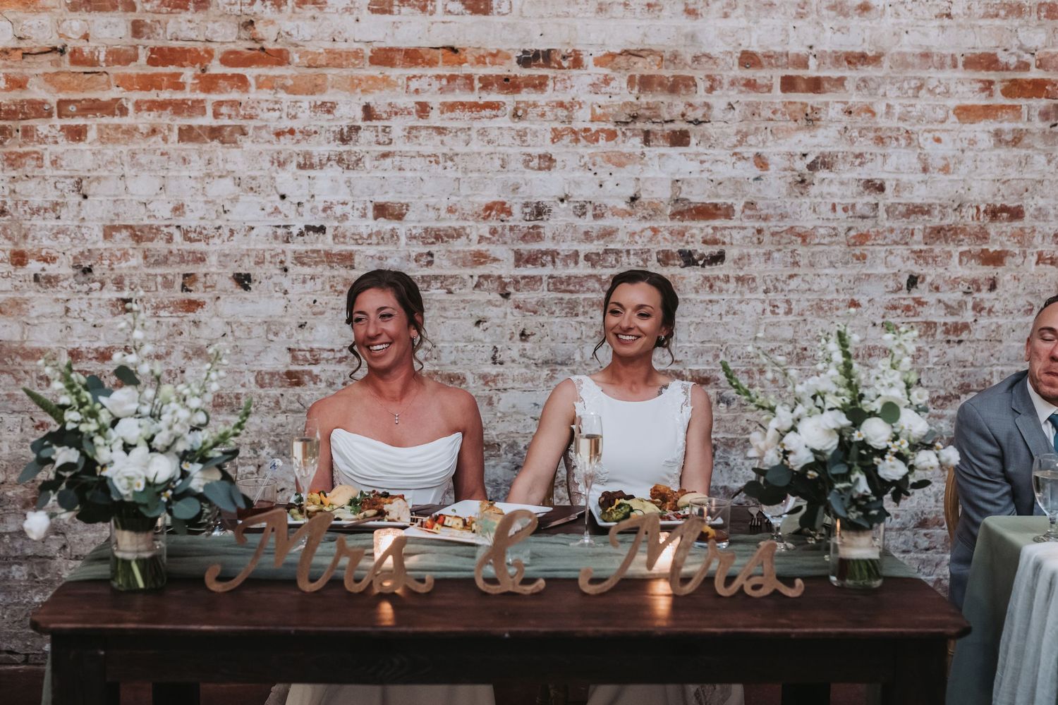 Two brides laughing joyfully at sweetheart table with Mrs and Mrs sign and white floral arrangements.
