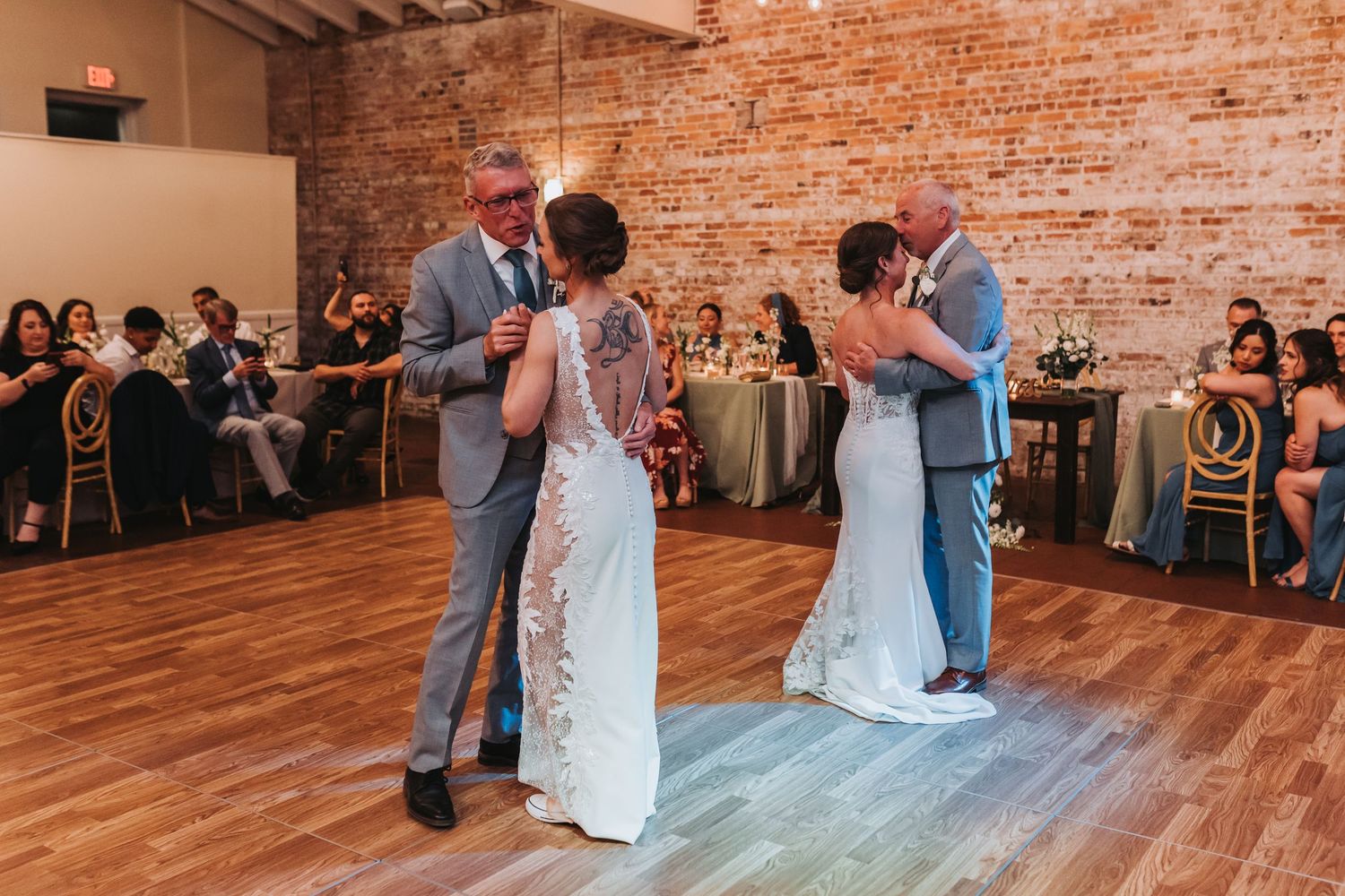 Two couples sharing first dance on wooden floor at elegant wedding reception with exposed brick walls.