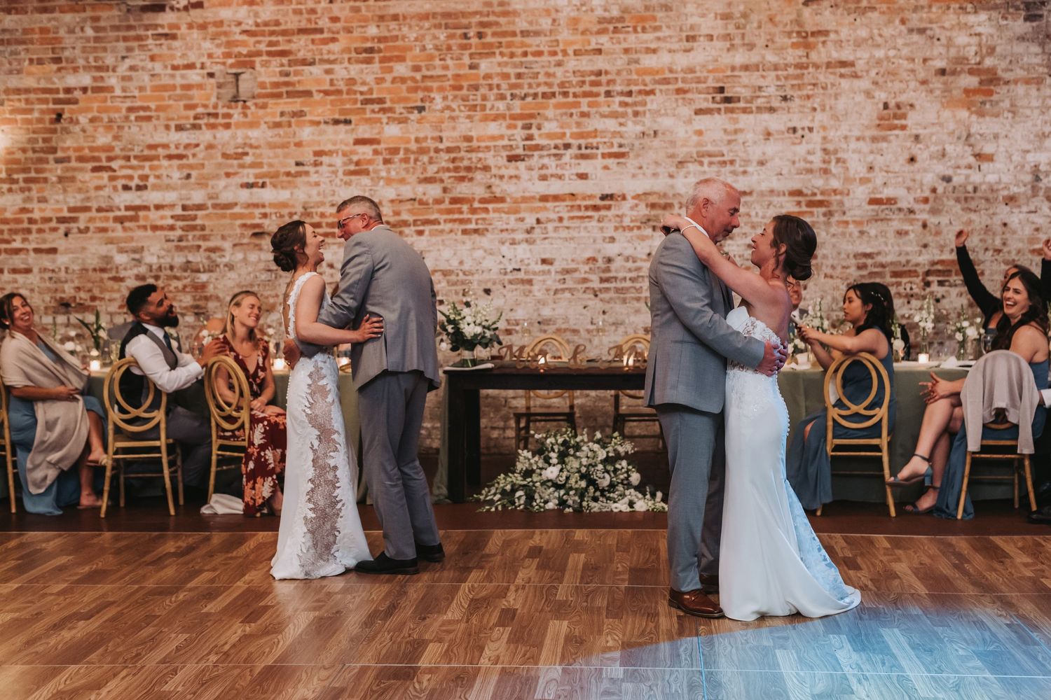 Two brides dancing with family members during parent dances at elegant brick-walled wedding reception.