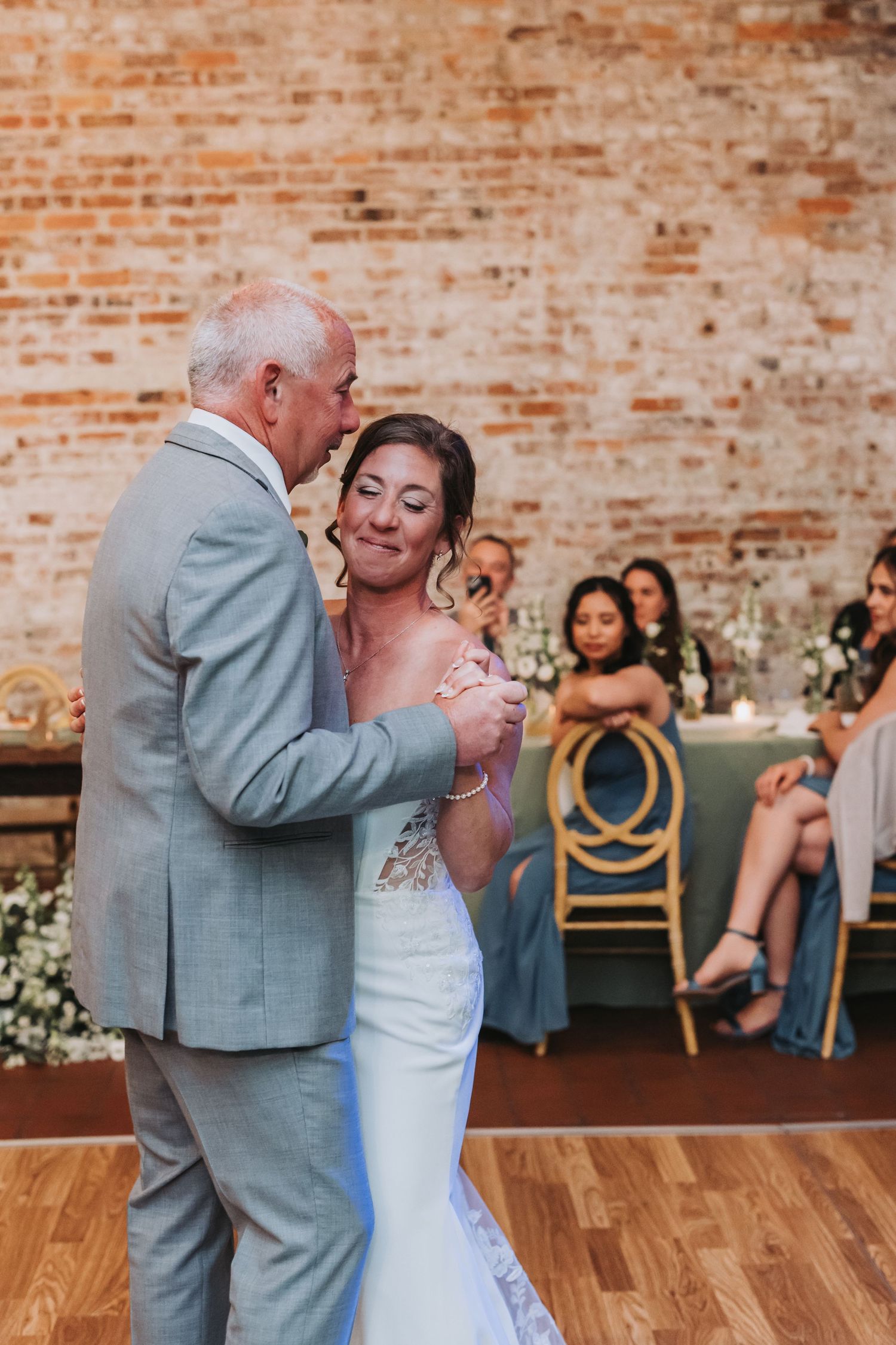 Emotional bride in lace wedding gown sharing a tender father-daughter dance in front of an exposed brick wall.