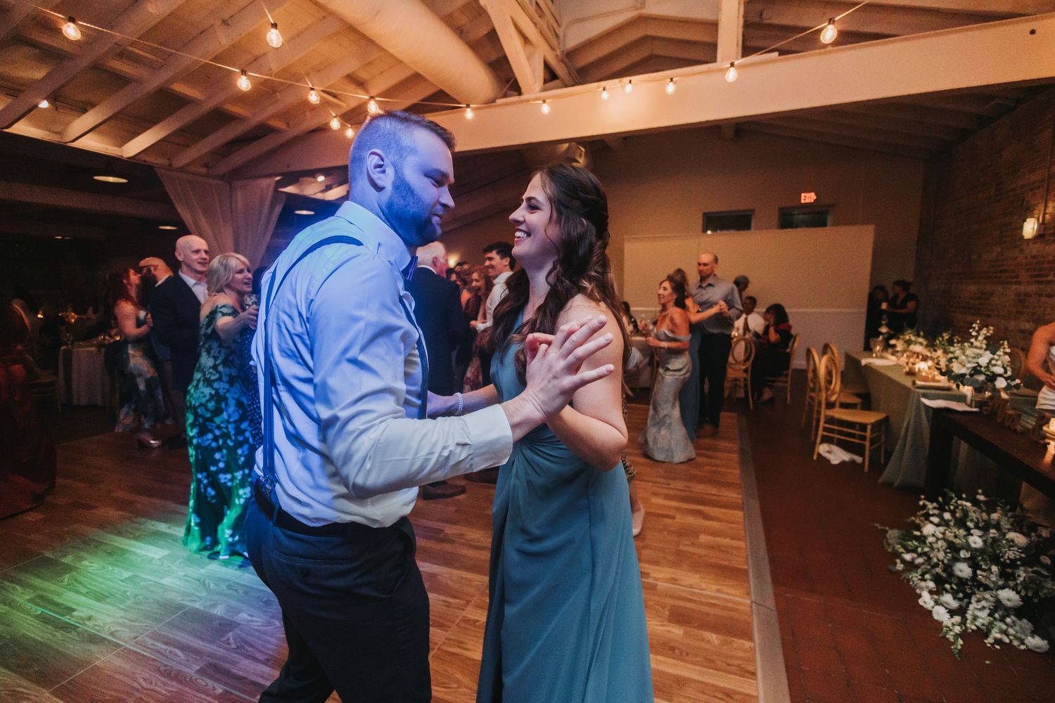 A couple dances joyfully together at a wedding reception with warm string lights overhead.