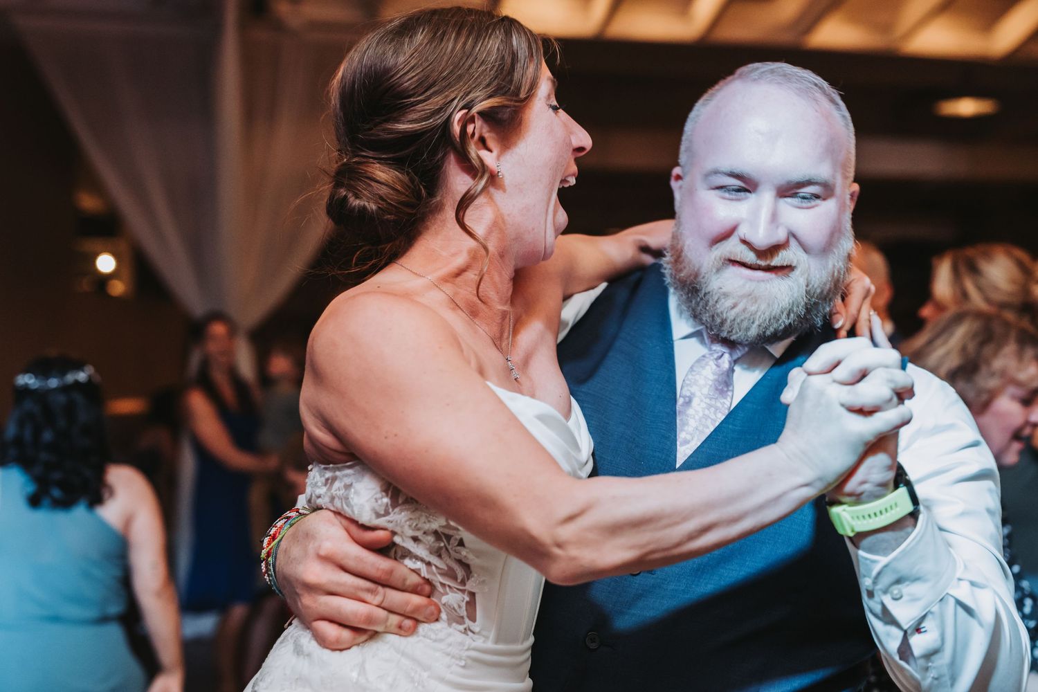 Bride in white gown laughing joyfully while dancing with bearded man in blue vest at wedding reception.