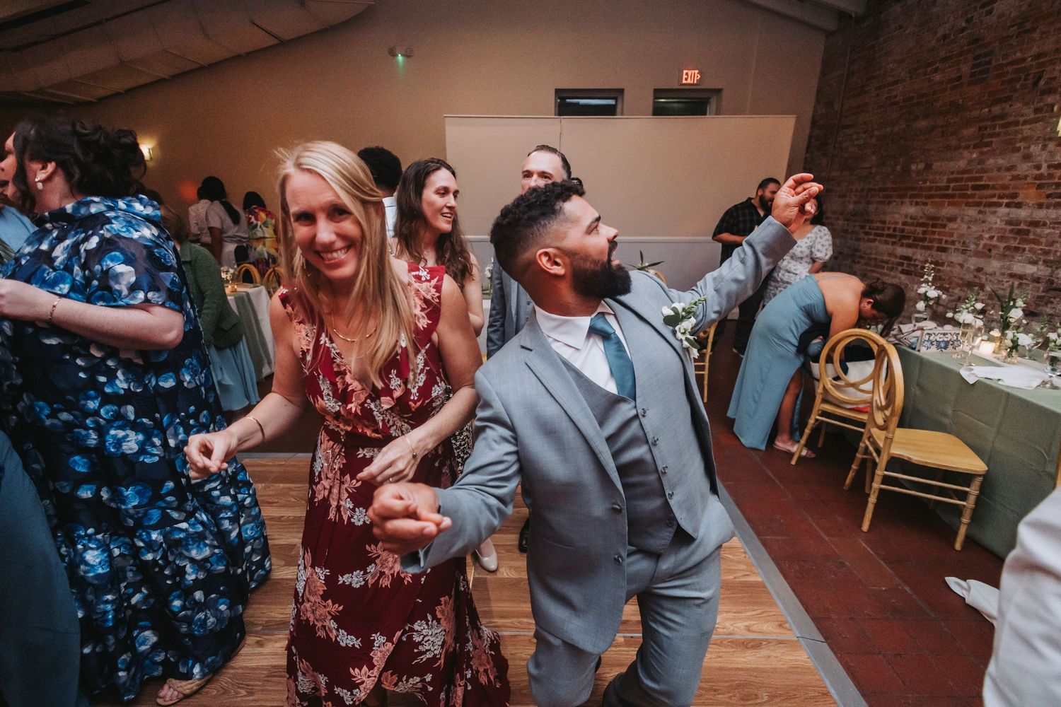 A bearded man in a gray suit dances energetically with a woman in a floral dress at a wedding.