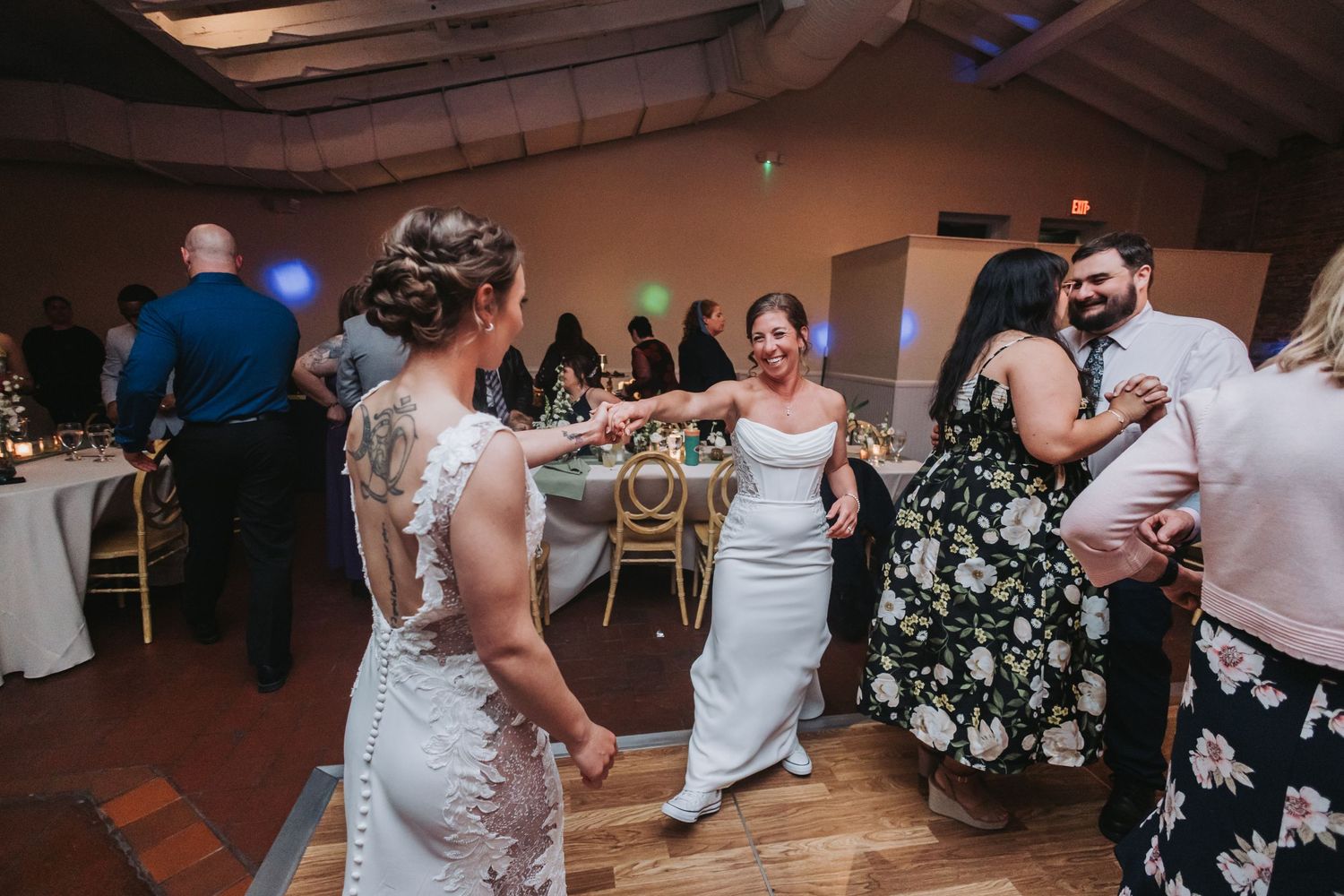 Two brides in white gowns dance together with guests at their wedding reception.