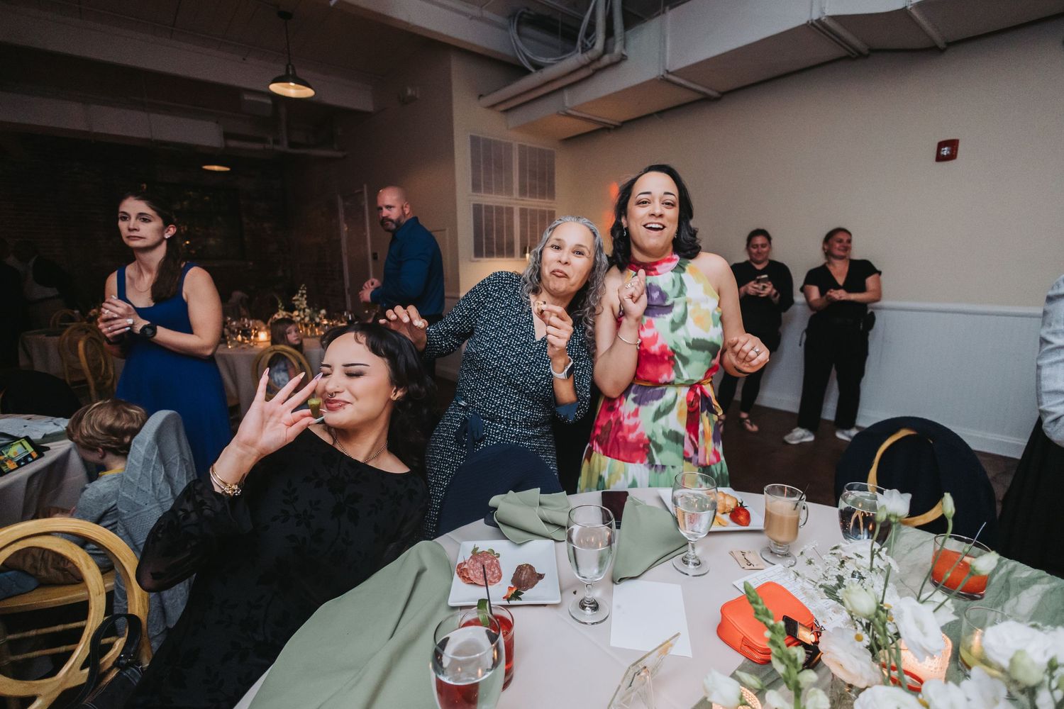 Women celebrating at a wedding reception dinner table, laughing and posing for photos in an elegant banquet hall.