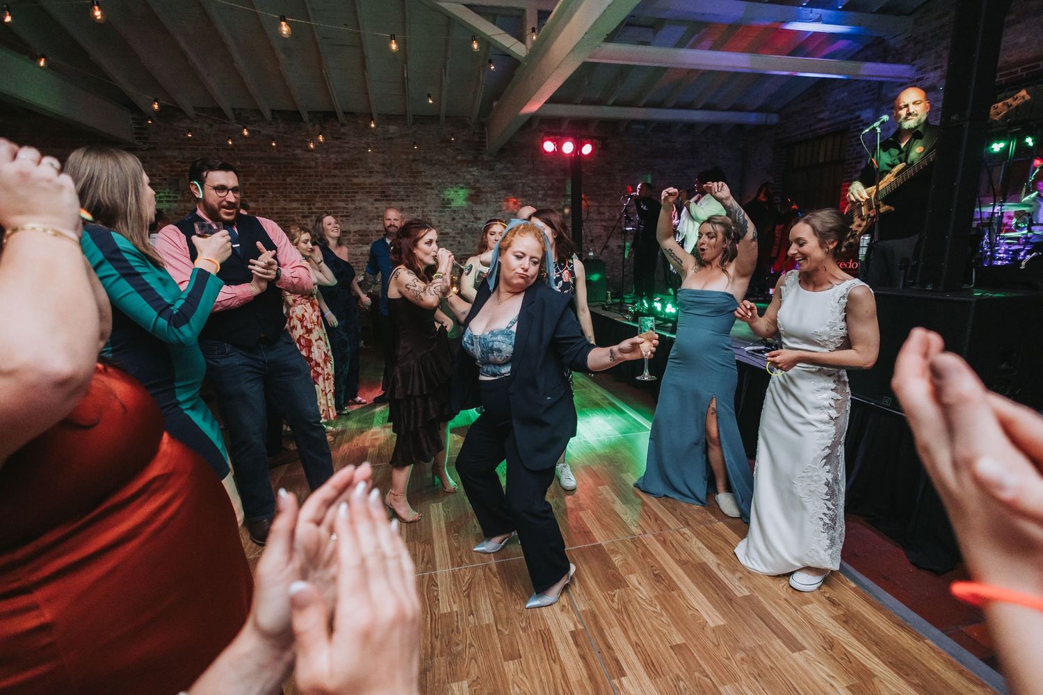 Wedding guests dance enthusiastically on a wooden dance floor with colorful stage lighting.