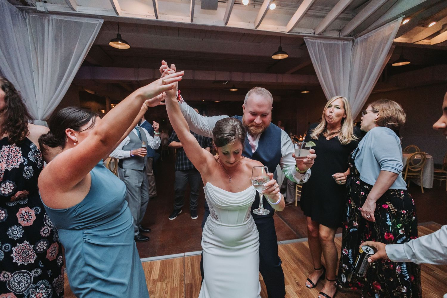 A bride in a white gown is twirled by guests on the dance floor at a lively wedding reception.