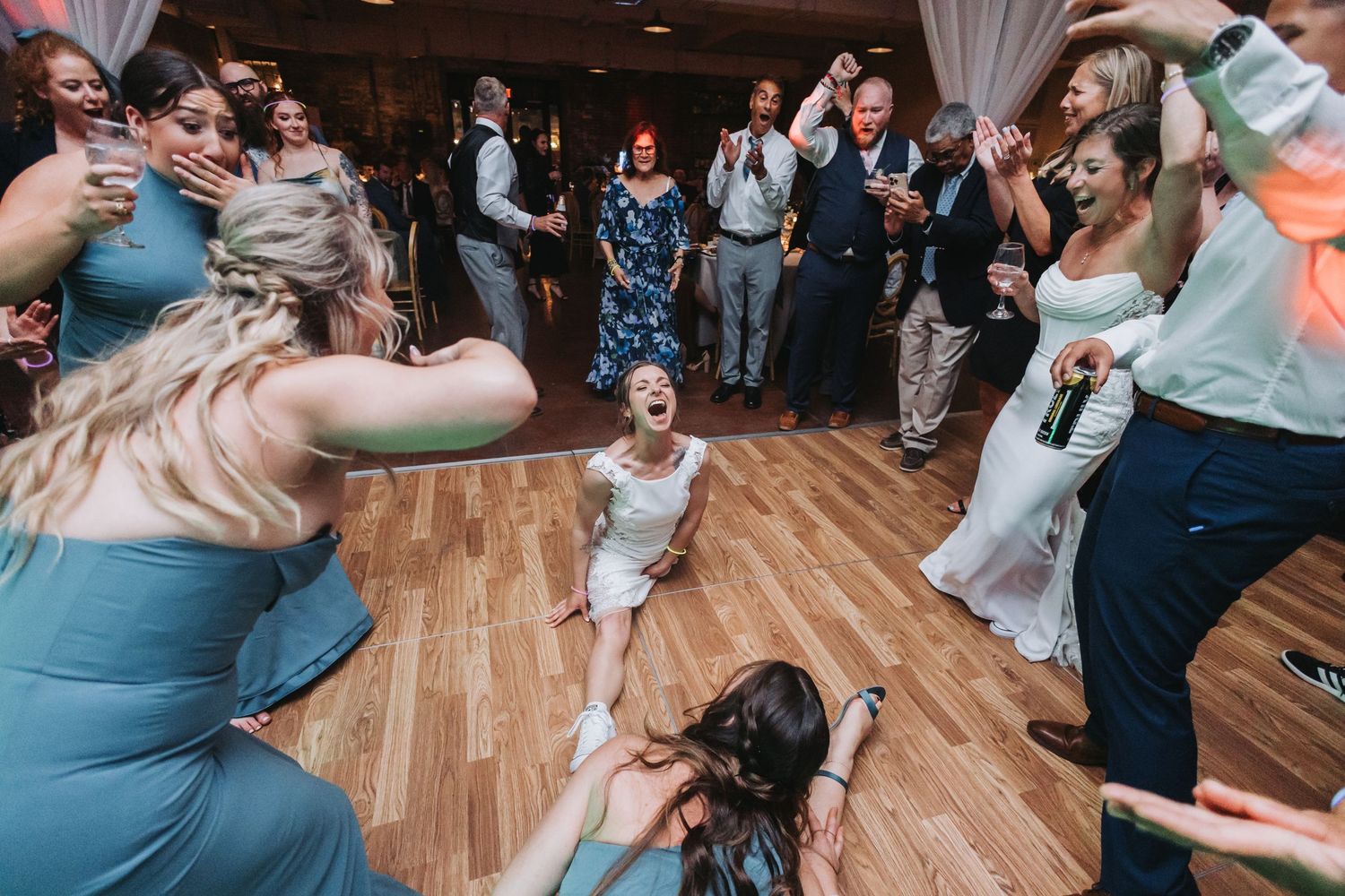 Bride laughing on dance floor surrounded by bridesmaids in blue dresses during lively wedding reception celebration.