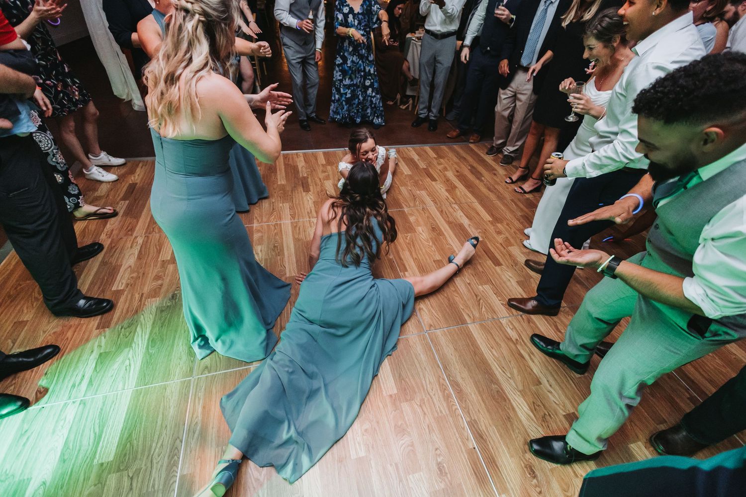 Wedding guests dancing energetically on wooden dance floor, woman in blue dress sliding dramatically at lively reception party.
