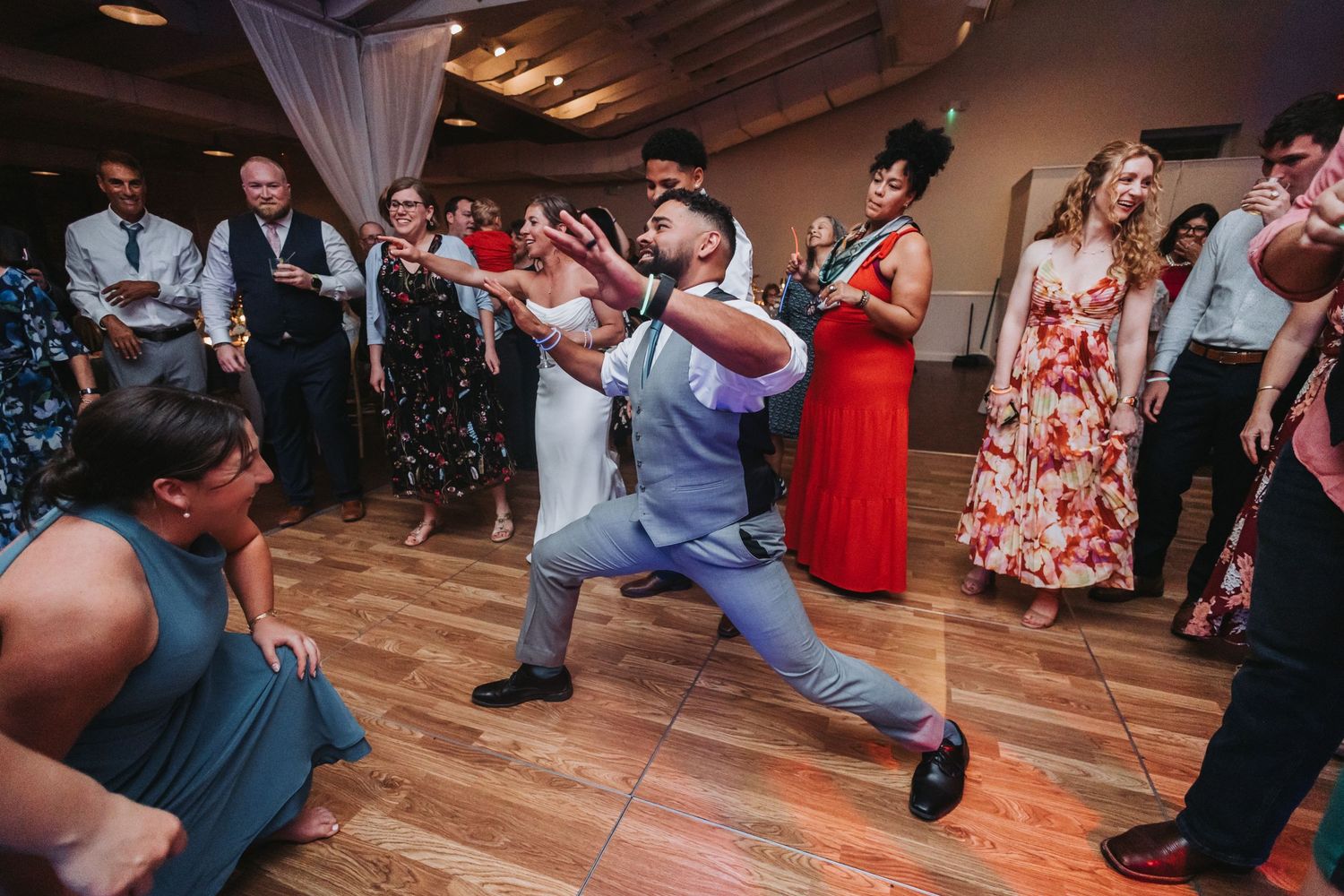 Guests dancing energetically at a wedding reception on a wooden dance floor in a decorated venue.