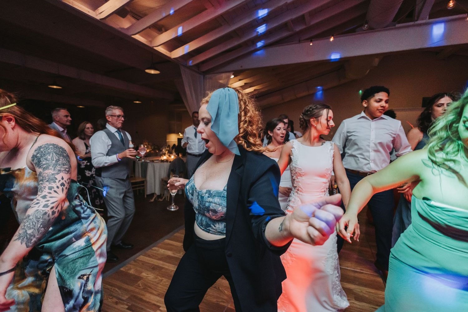 Excited wedding guests dancing at reception, woman with blue hair wearing mask celebrating on colorful lit dance floor.