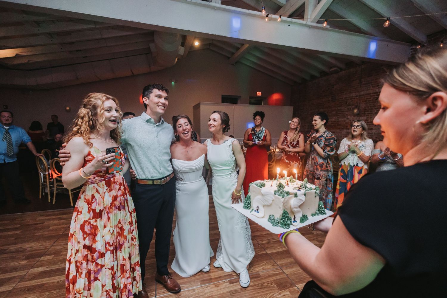 Wedding guests smile as a woman presents a white celebration cake with candles at a rustic venue reception.