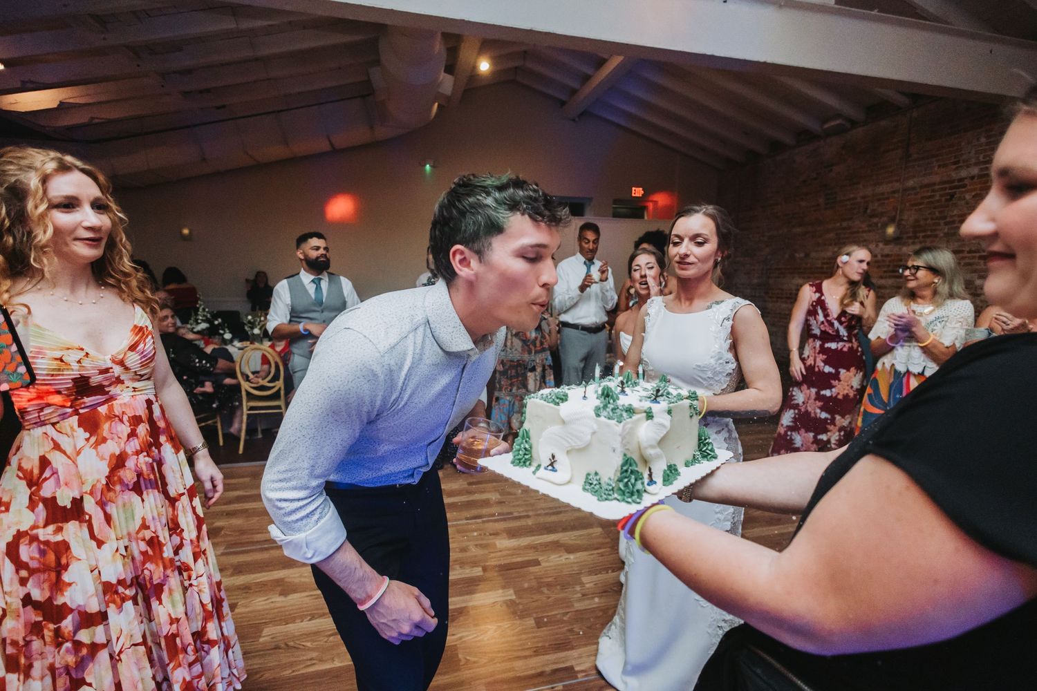Groom blowing out candles on white wedding cake held by guests at elegant brick-walled reception venue.