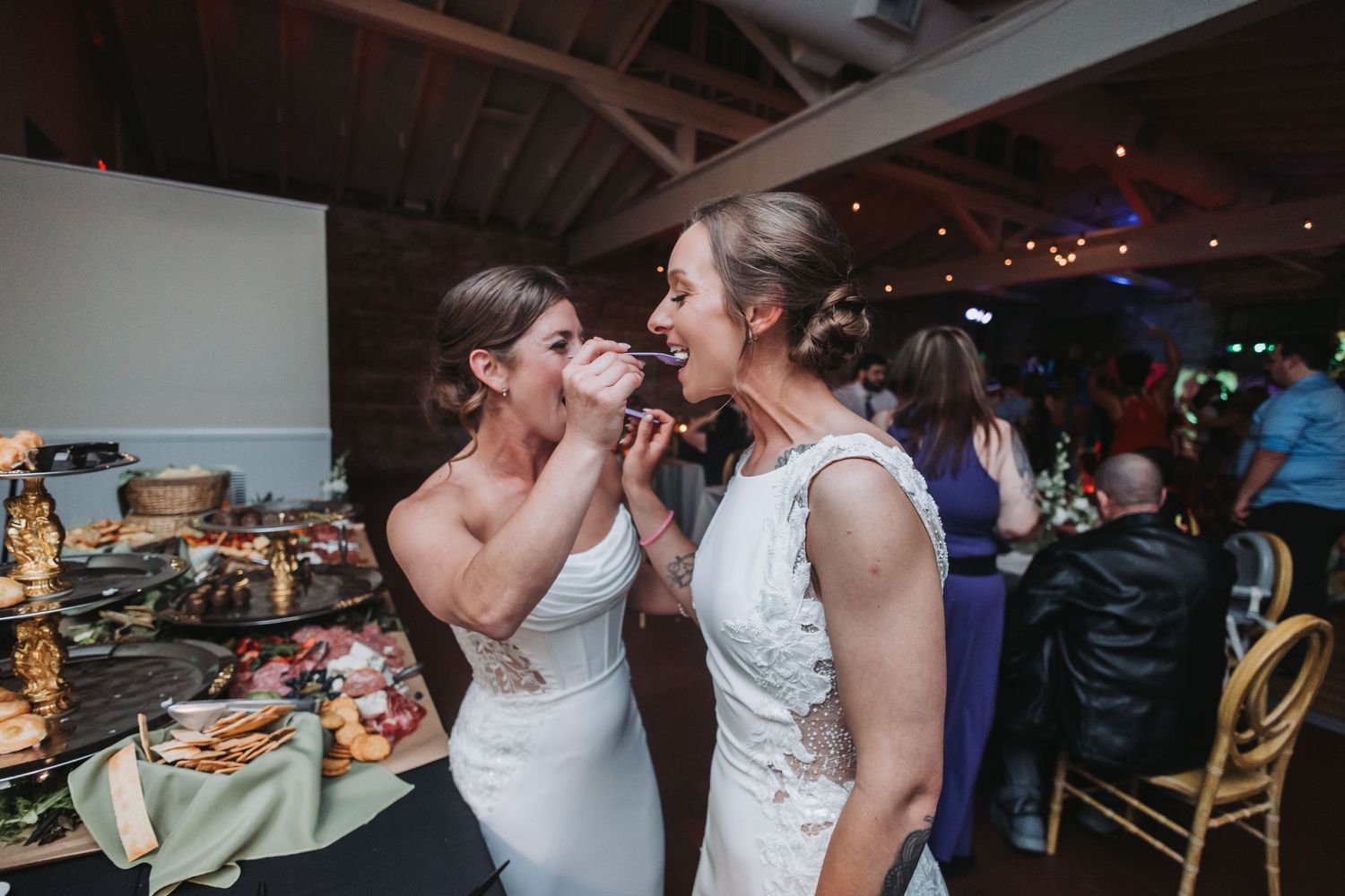 Two brides in white dresses share a joyful moment eating together at their wedding reception.