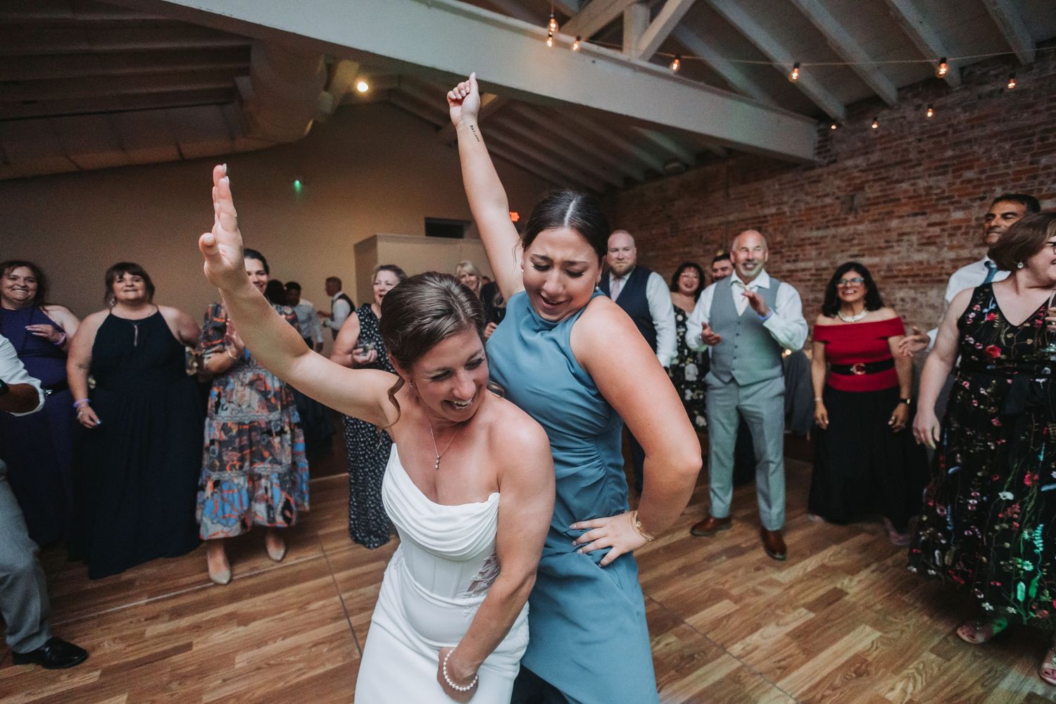 Bride and bridesmaid dancing energetically at a wedding reception with guests watching.