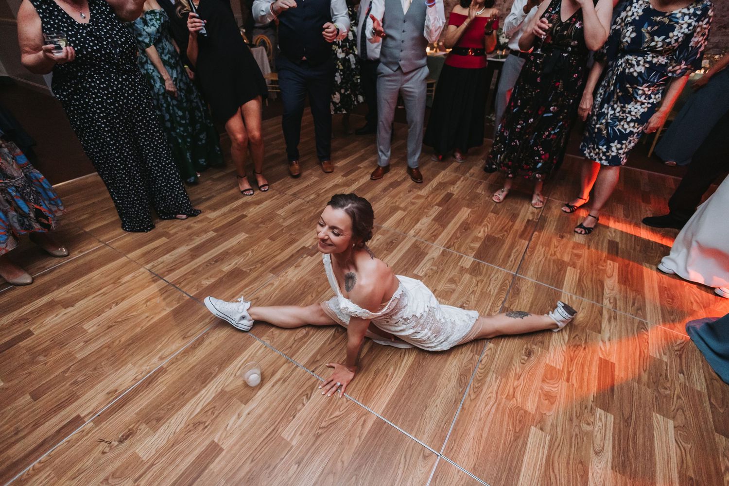 Woman doing the splits on a wooden dance floor at a wedding reception with guests watching.