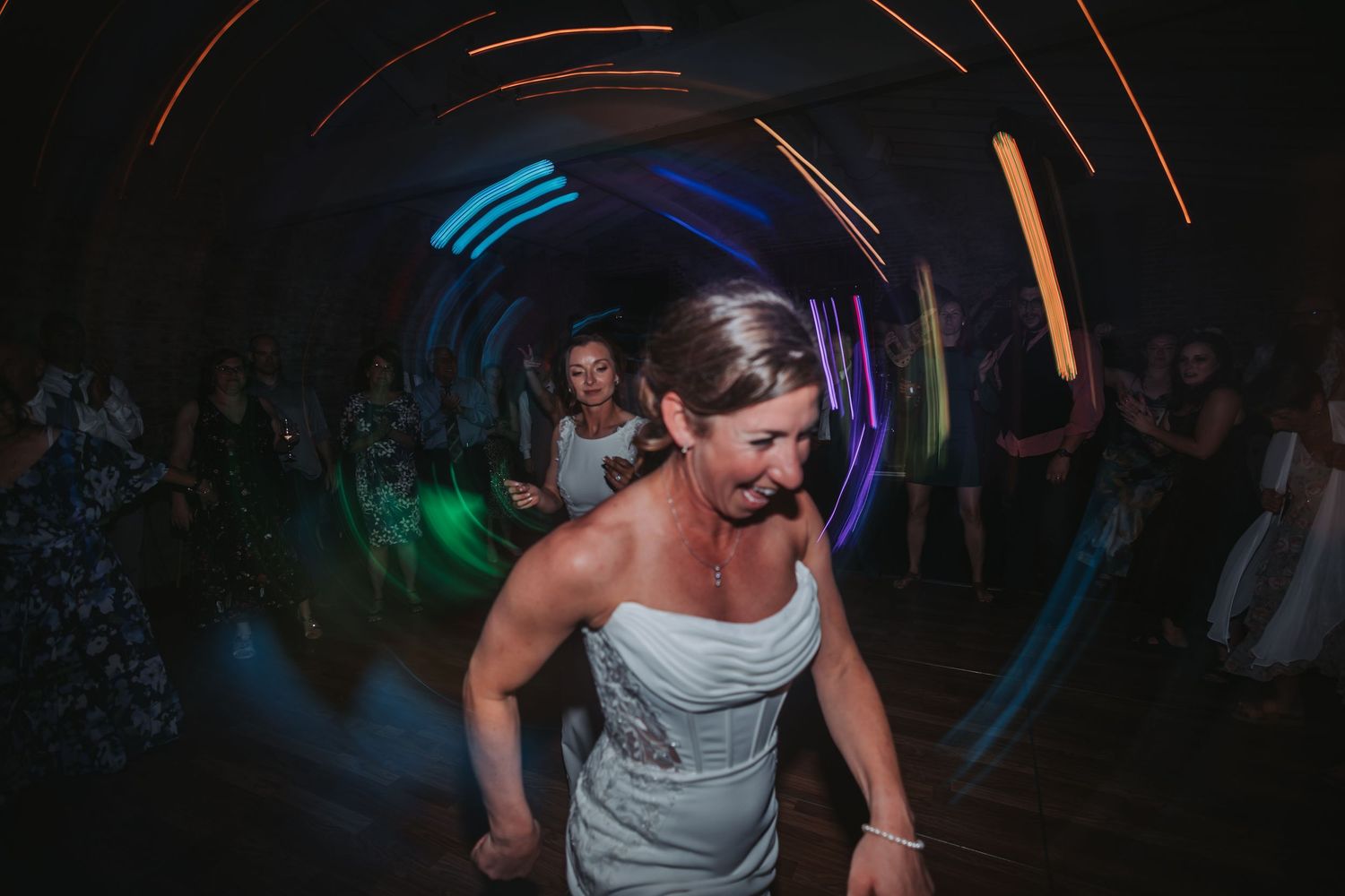 Bride in white gown dancing joyfully under colorful light trails at wedding reception.