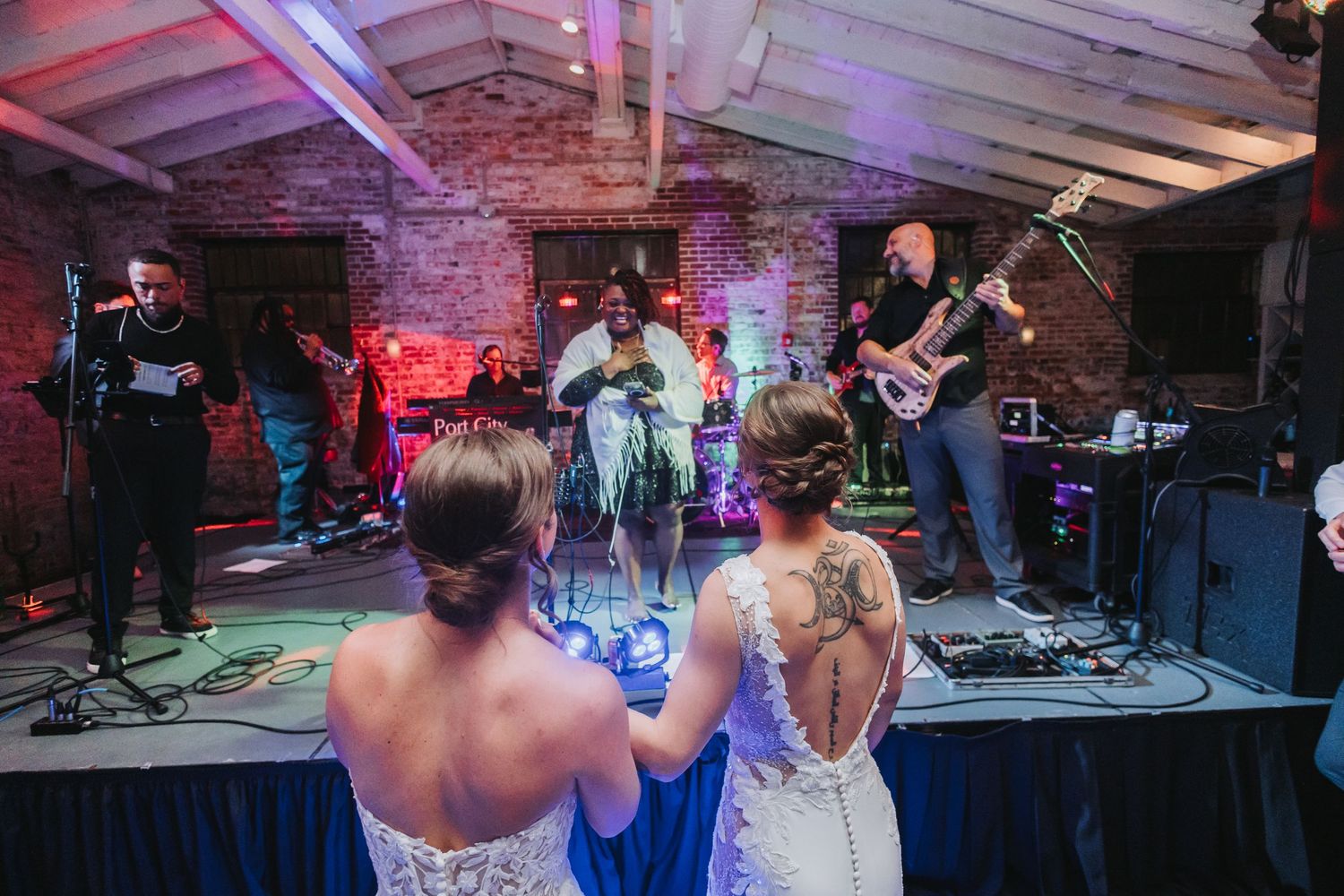Two brides in white gowns watching a live band perform at their wedding reception.