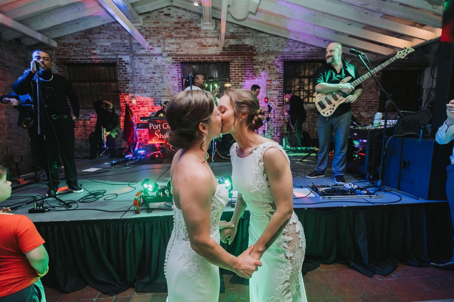 Two brides share a kiss on the dance floor with a live band performing in the background at their wedding reception.