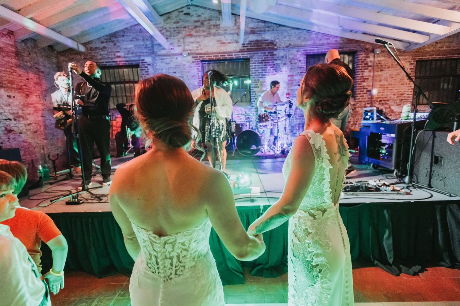 Two brides in white lace gowns hold hands facing a live wedding band performing on stage.