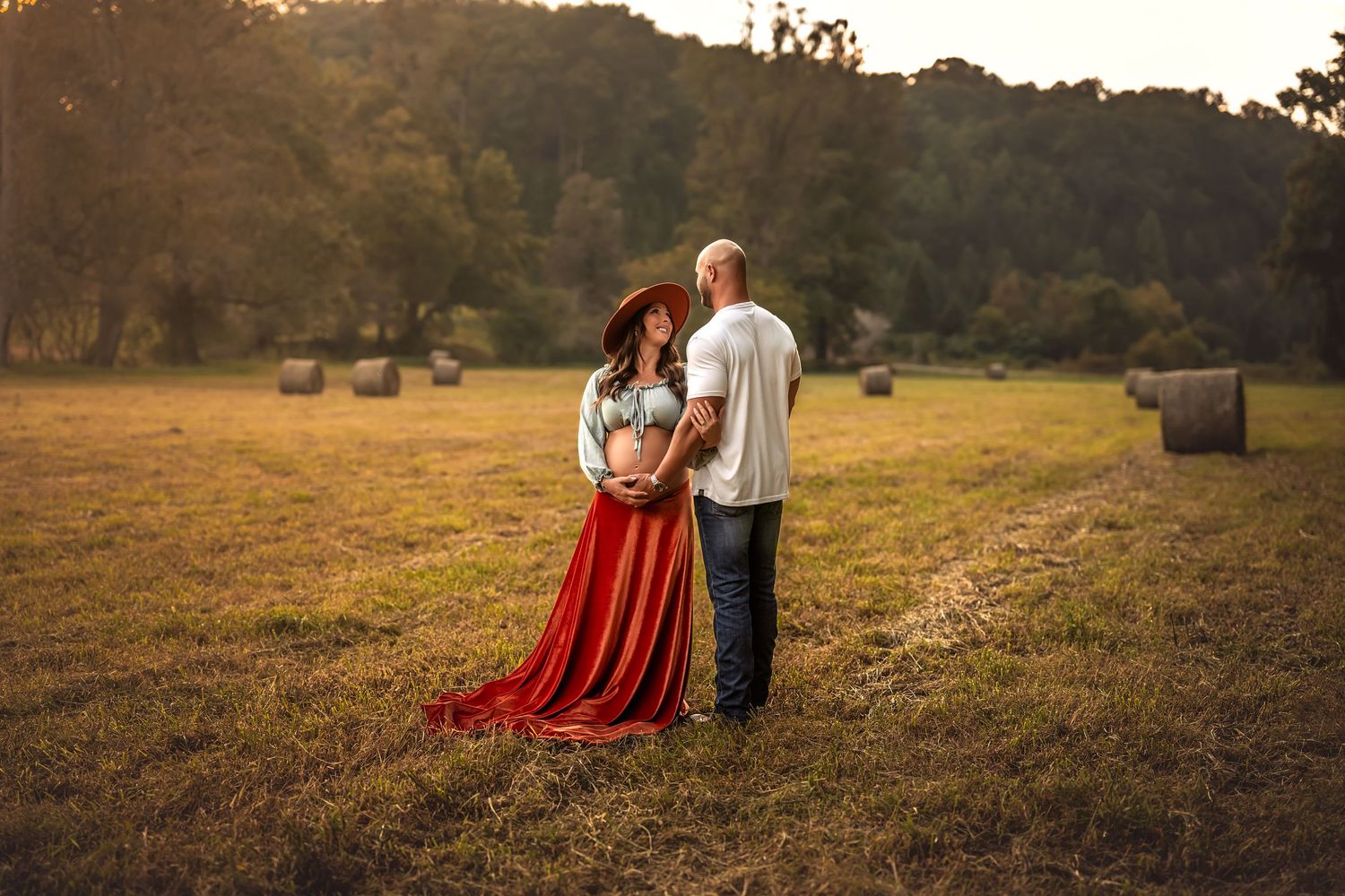 Pregnant couple embracing in golden hour field with hay bales, woman in red flowing skirt and sun hat.