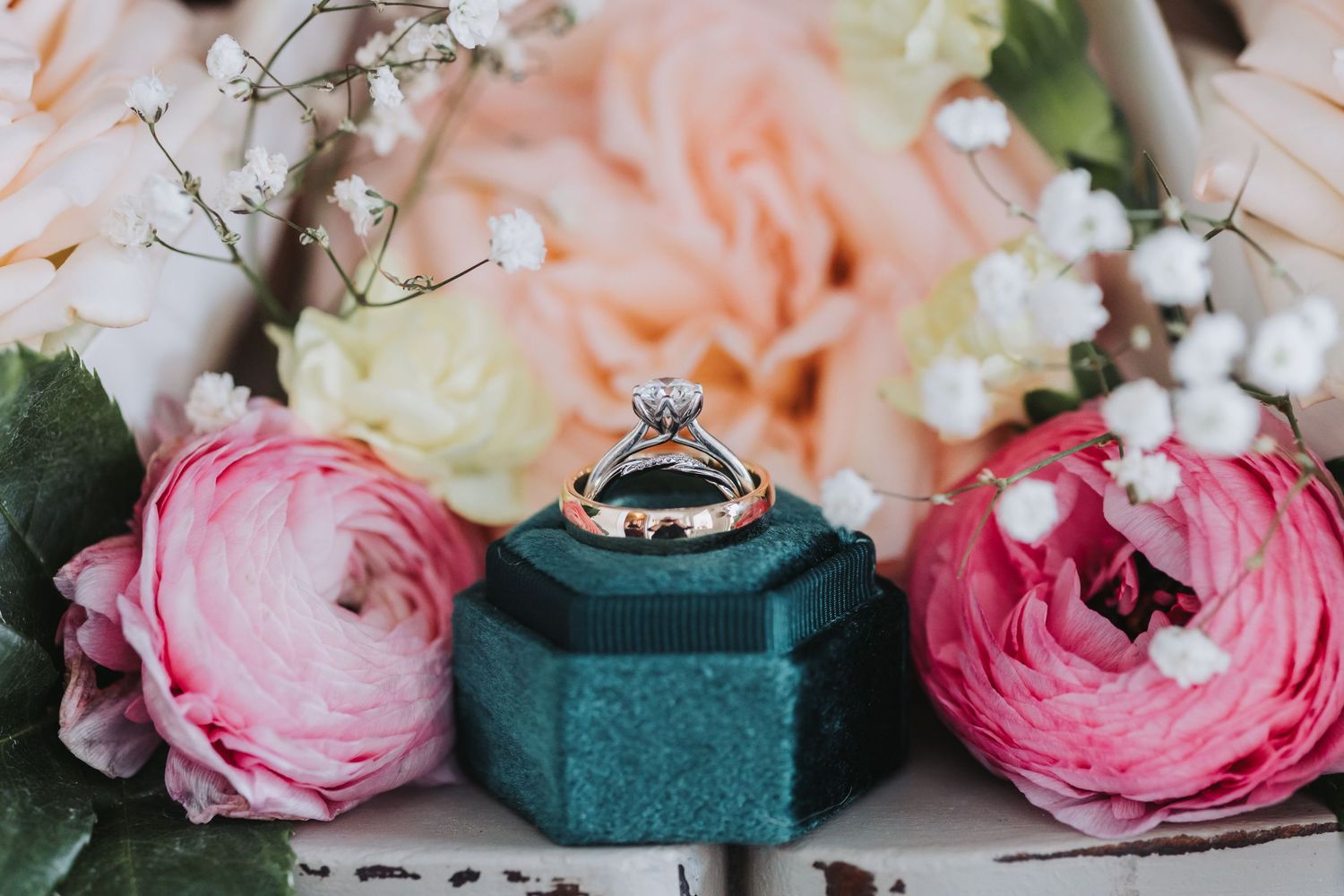 Diamond engagement ring displayed on teal velvet box surrounded by pink ranunculus and baby's breath flowers.