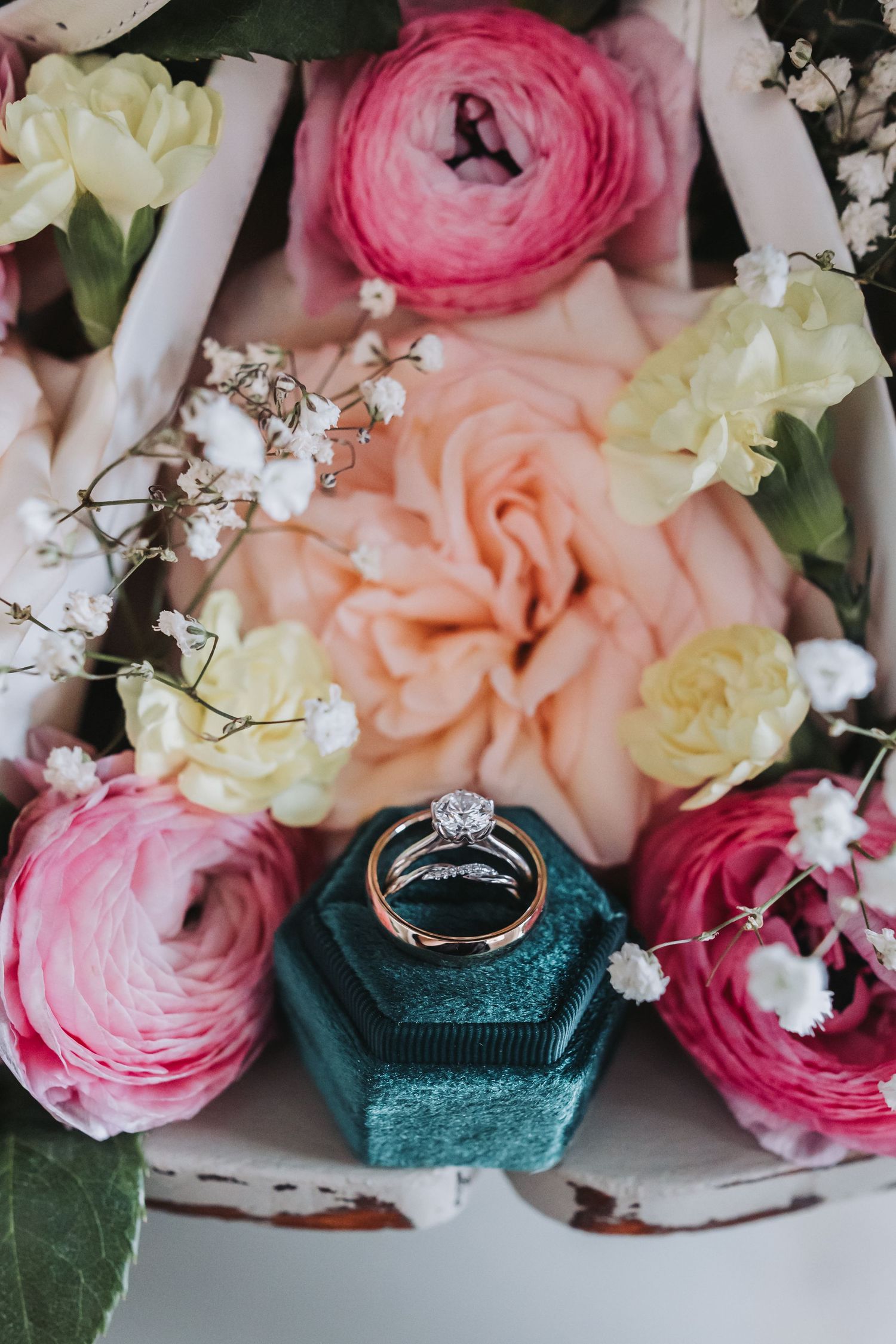 Diamond engagement ring and gold wedding band in teal velvet box surrounded by pink and cream flowers.