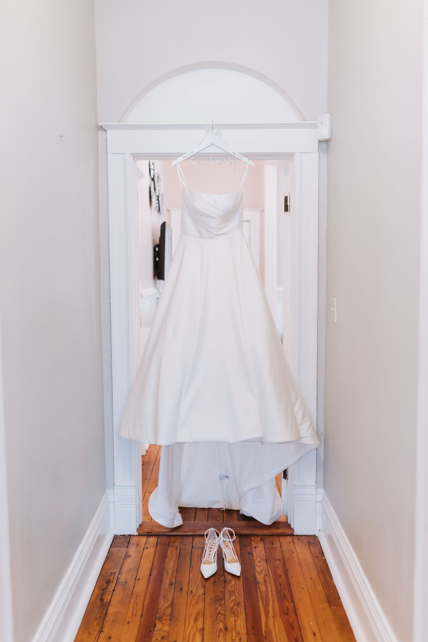 Elegant white strapless wedding gown hanging in doorway with bridal heels below.